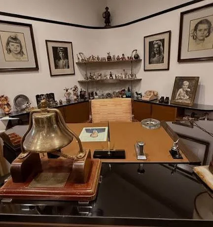 Sala de oficina con escritorio de madera, lámpara antigua, fotos enmarcadas de niños, y estanterías con figuras y objetos decorativos.