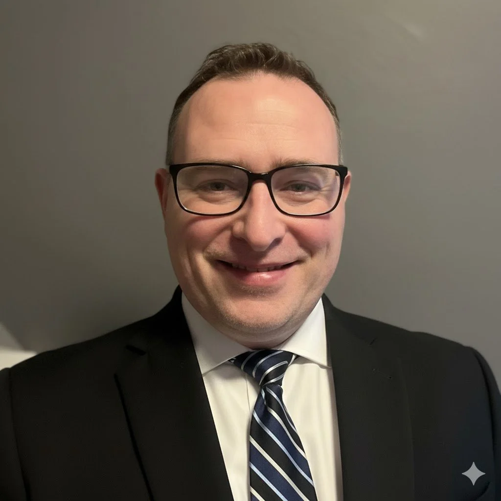 A man with glasses smiling, wearing a black suit, white shirt, and a striped tie, against a plain gray background.