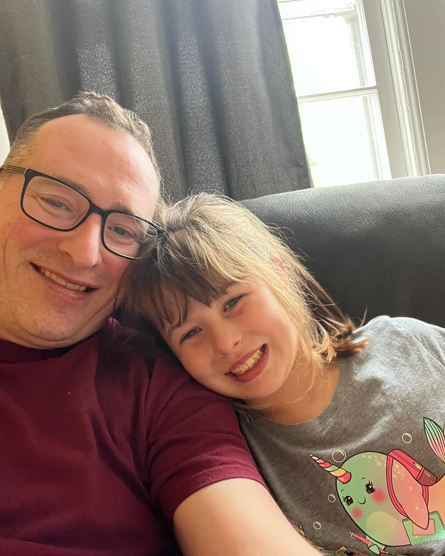A man and a young girl smiling and taking a selfie together indoors, with a window and curtains behind them.