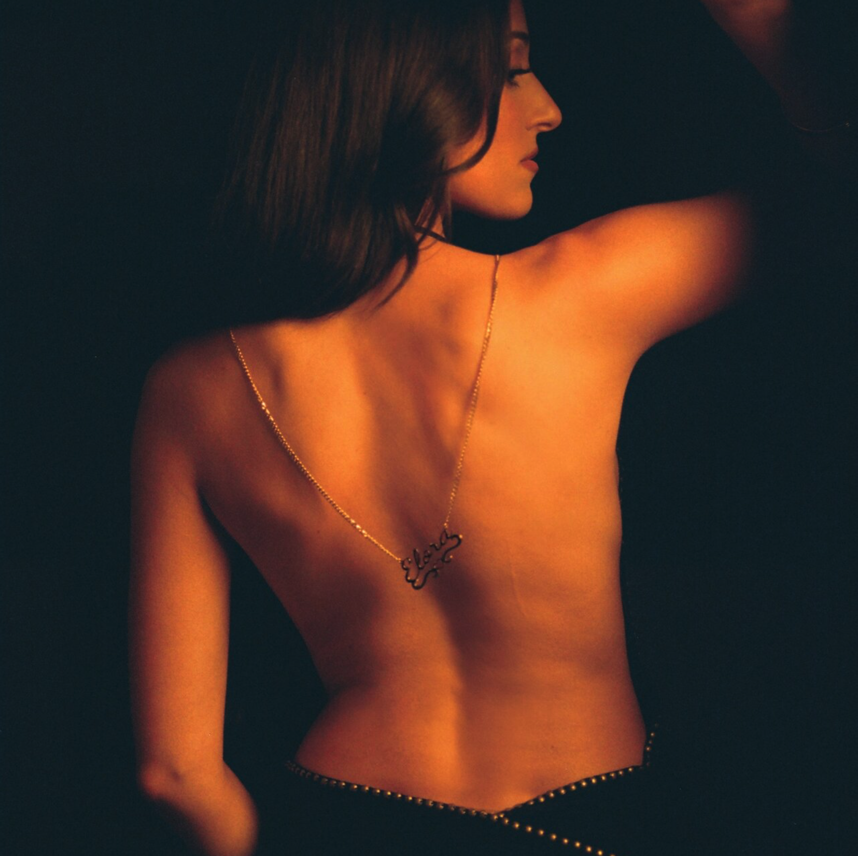 A woman with dark hair and fair skin, topless, facing sideways, with her left arm raised. She is wearing a gold necklace with a name or word in cursive, and a gold chain around her waist. Elle Eliades - Elora