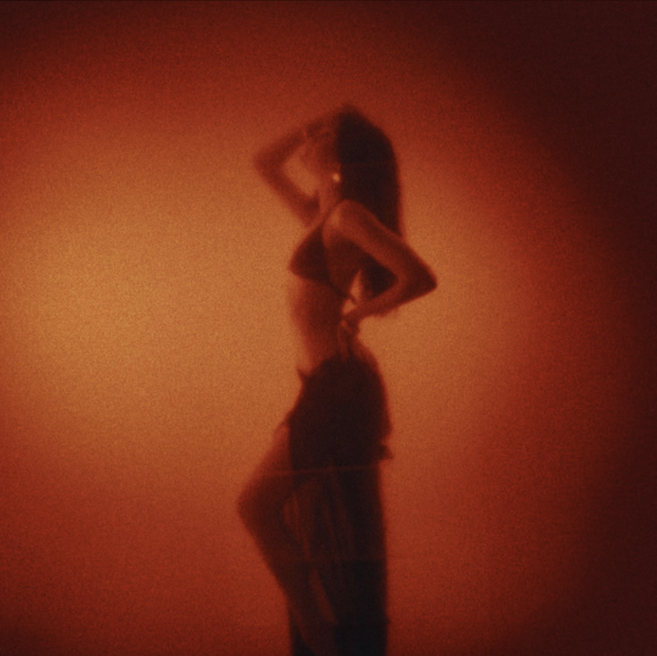 A woman with long dark hair posing against a reddish-orange gradient background, wearing a black top, patterned pants, and lifting one arm above her head. Julia Lostrom