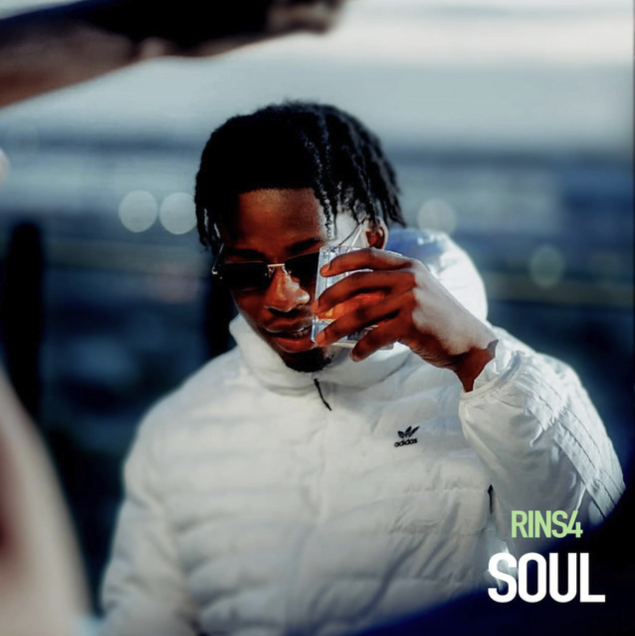 A man with dreadlocks wearing sunglasses and a white Adidas jacket, holding a glass of what appears to be whiskey, standing near the water with blurred lights in the background, with the words 'RINS4 SOUL' in the bottom right corner.