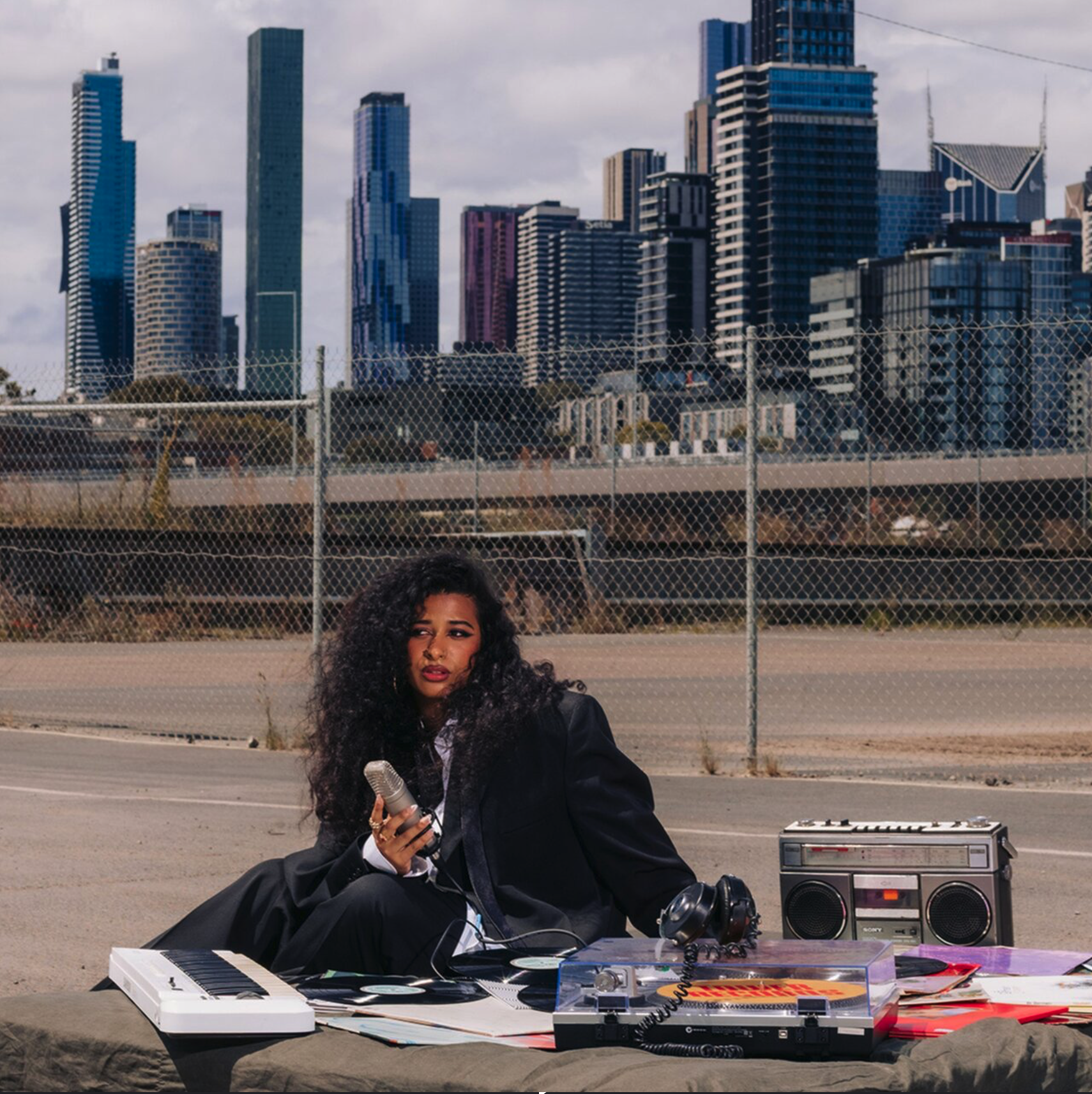 A woman with curly hair, dressed in a dark blazer and sitting on the ground, holds a microphone. She is surrounded by music equipment, including a keyboard, a boombox, a vinyl record player. Shreta EP