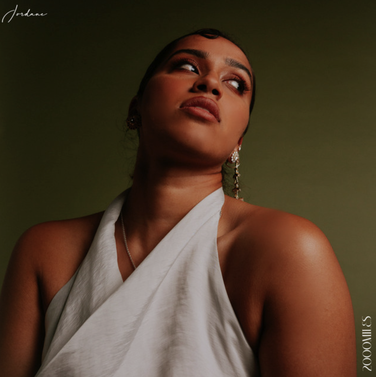 A woman with dark hair and large earrings, wearing a white halter top, looking upwards against a dark green background. Jordane