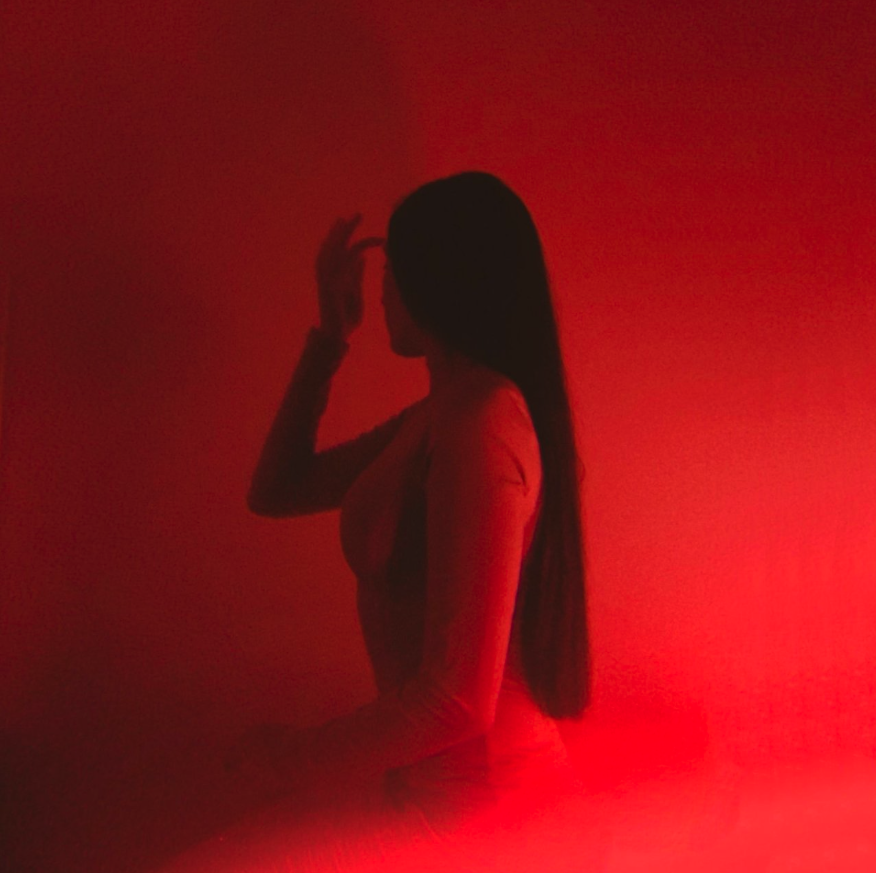 Silhouette of a woman with long hair, standing against a red-lit background, touching her forehead with her right hand. Elle Eliades