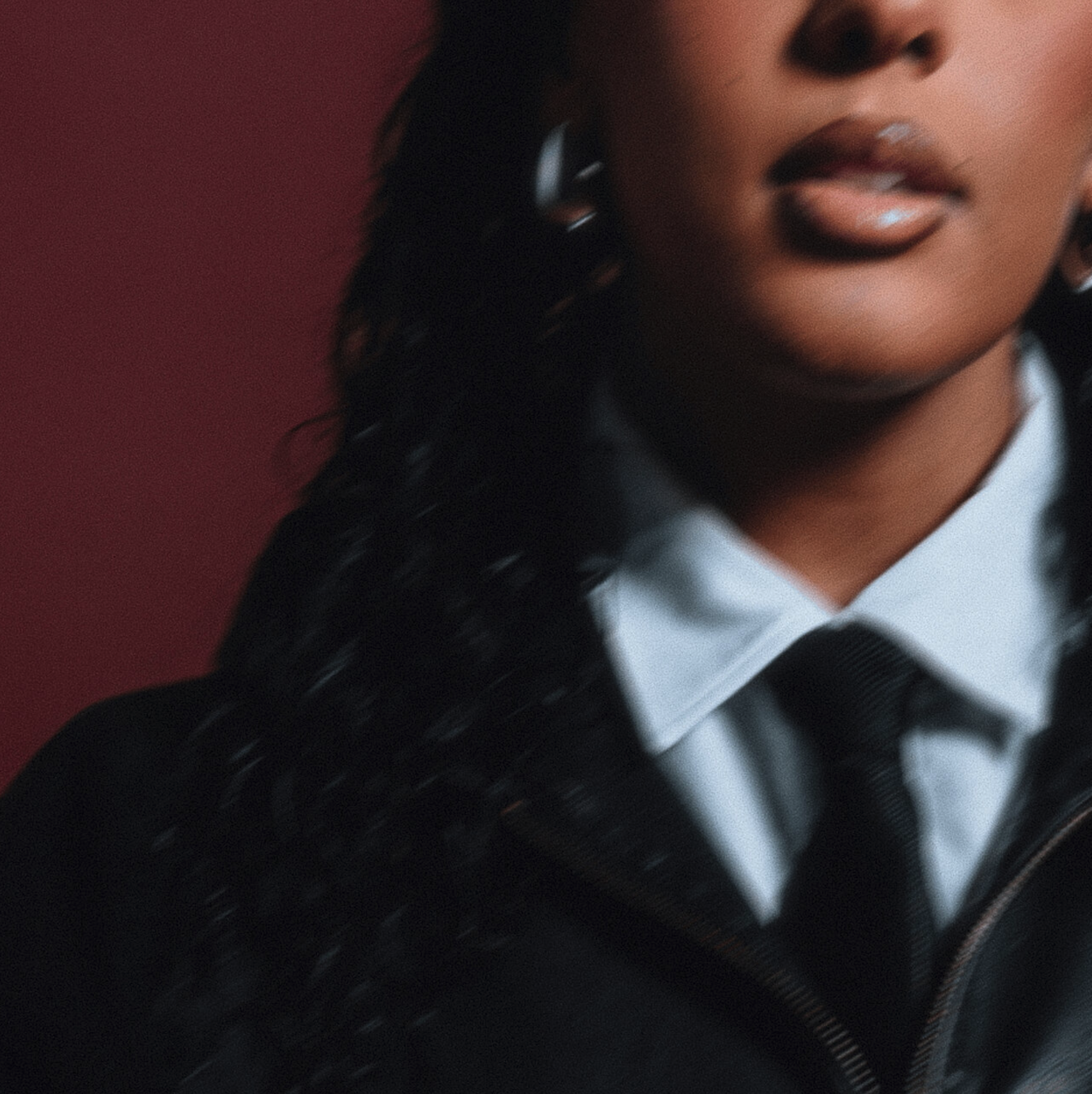 Close-up of a woman with curly black hair, wearing a white collared shirt, black tie, and black jacket, against a dark red background. Soli, Obvious