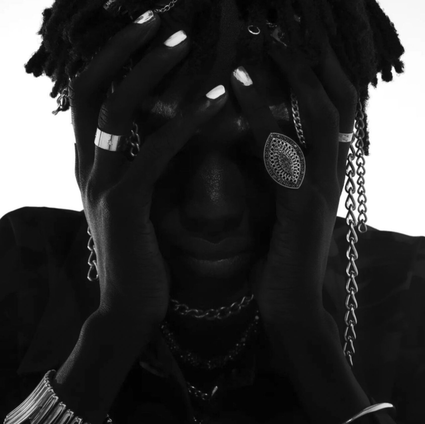 A person with dark skin and dreadlocks covering their face, wearing multiple jewelry pieces including rings, chains, bracelets, and a large decorative pendant, posed in a black and white photo. Tebir Different