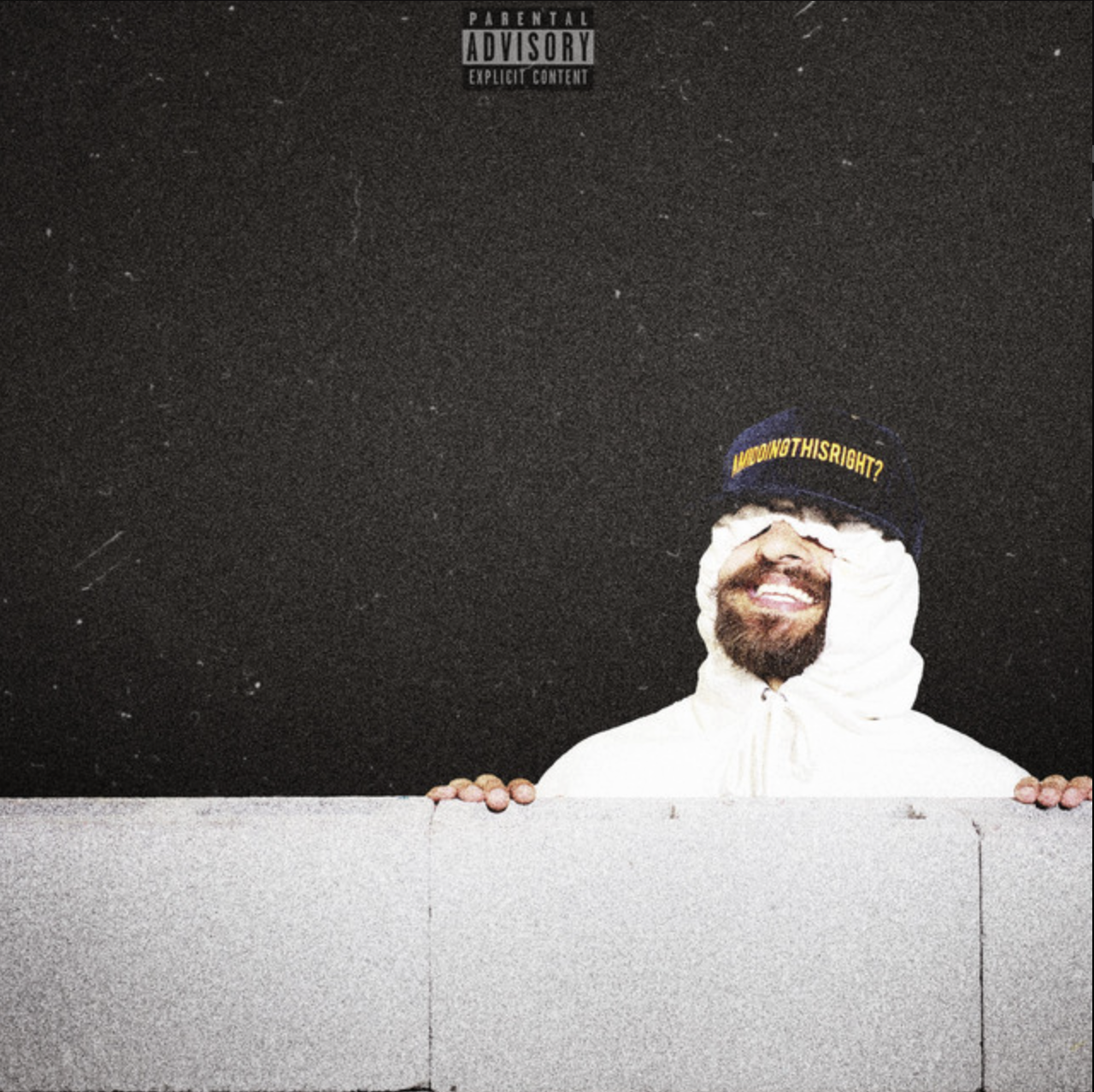 A man smiling, wearing a dark blue hat with yellow text and a white hoodie with the hood up, peeking over a concrete wall against a dark background. Sunny Roger