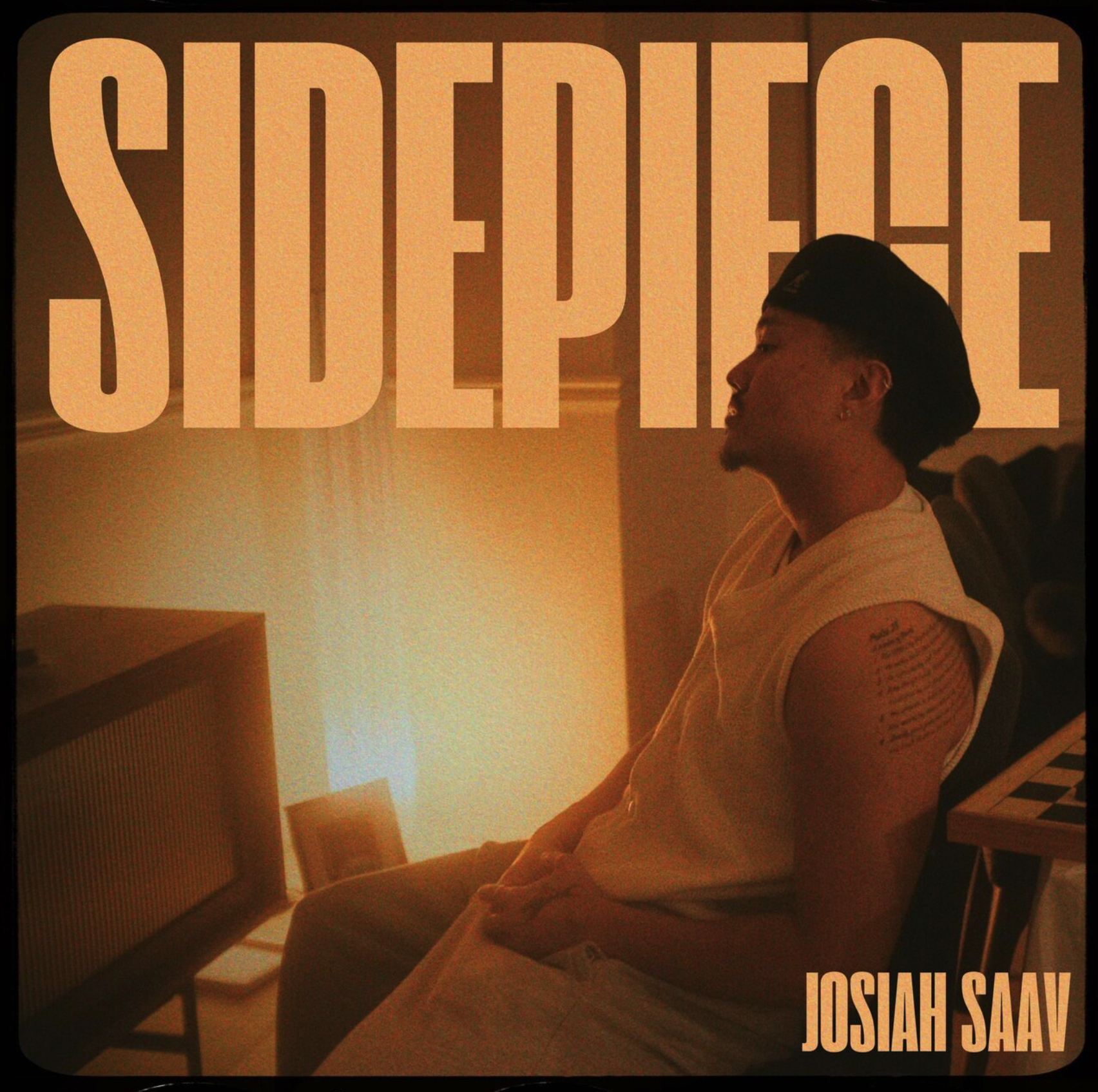 A young man with a tattoo on his upper arm, wearing a sleeveless shirt and a black cap, sitting with eyes closed in a dimly lit room, with a large word 'SIDEPIECE' overlaid at the top and 'JOSIAH SAAV' at the bottom right.