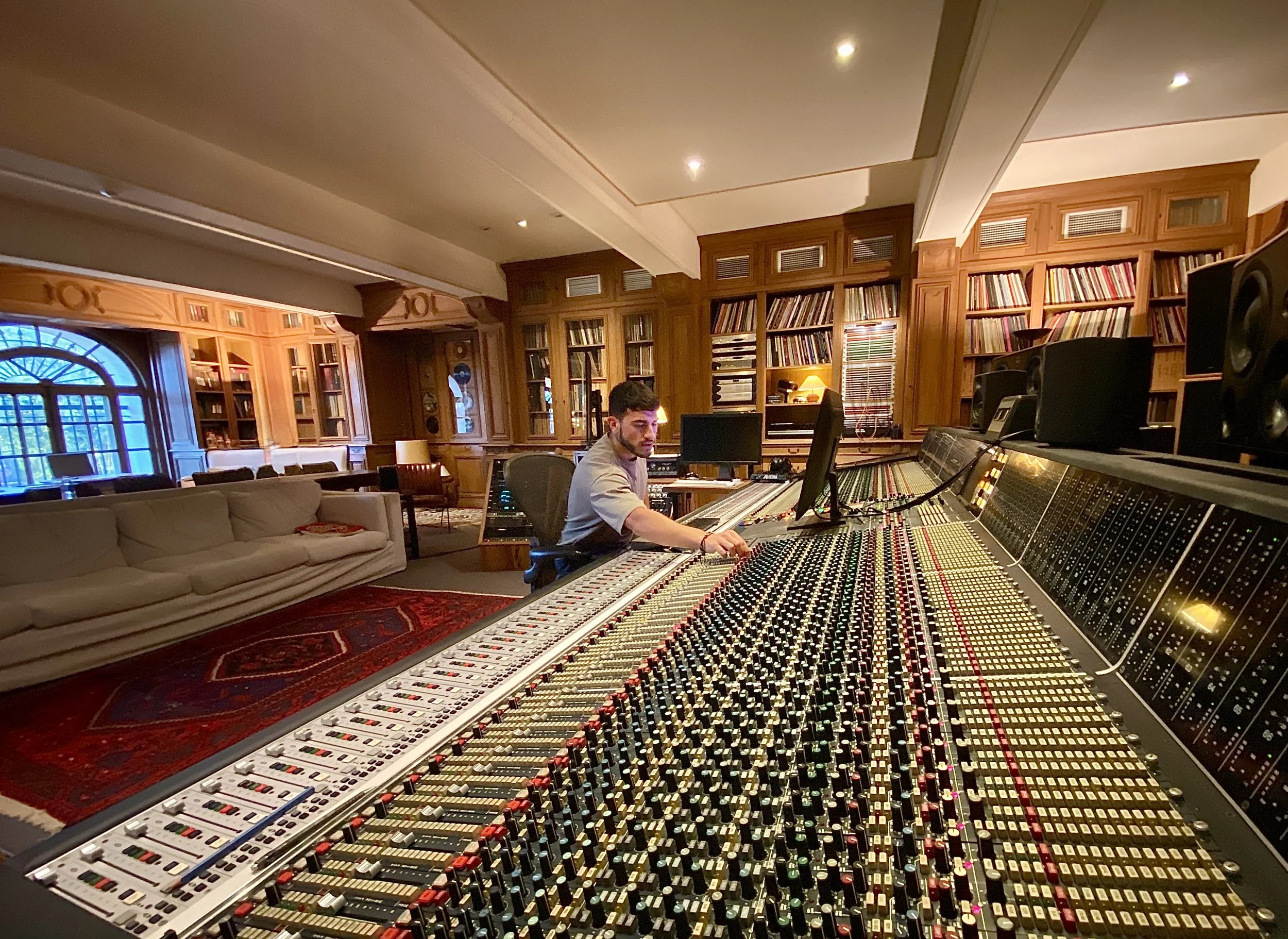 A man working at a large audio mixing console in a recording studio with wooden shelves filled with books in the background. Mix With The Masters. Studio in France