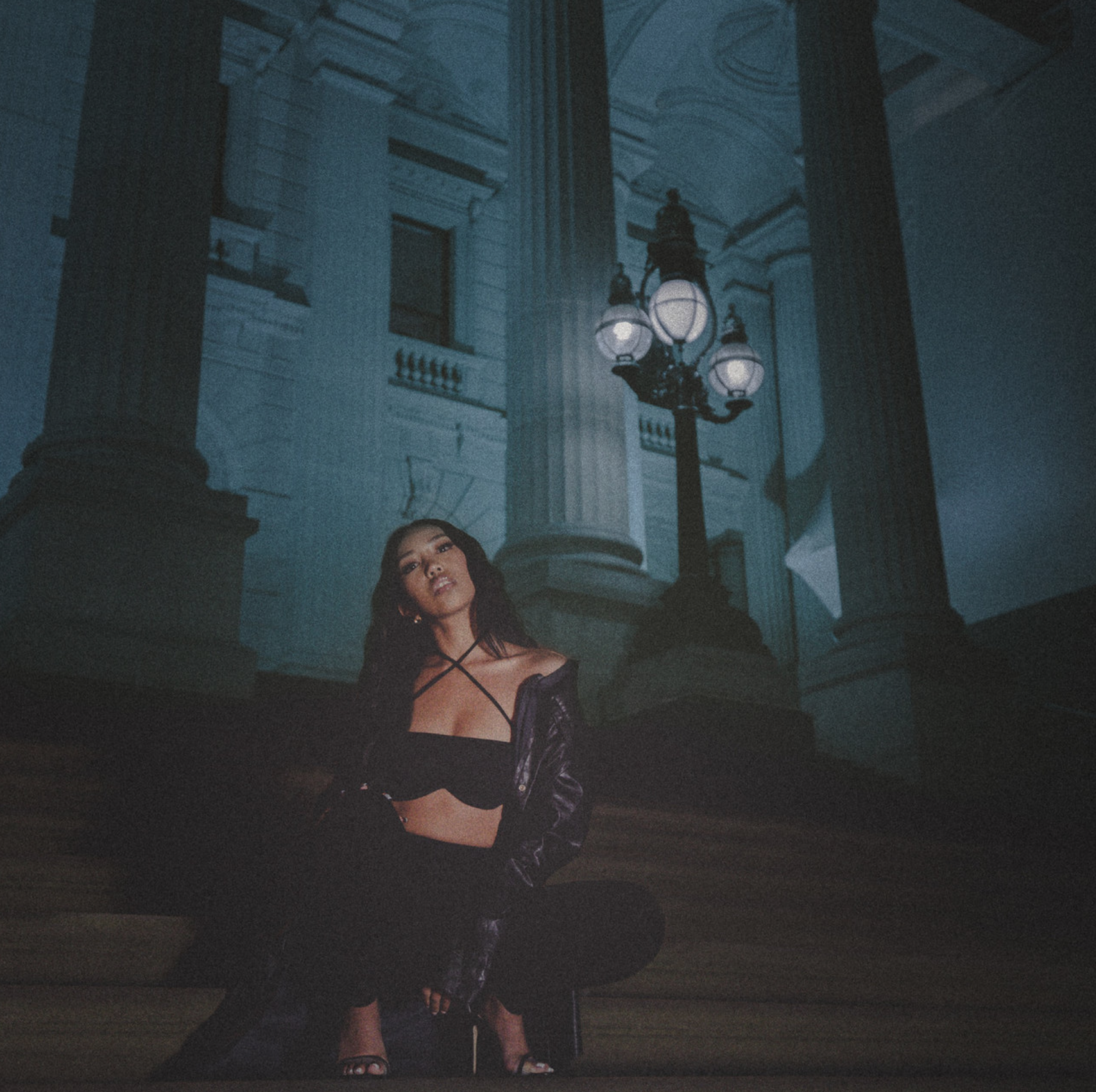 A woman with dark hair and dark eye makeup sitting on the steps of a grand building with large columns and an ornate lamp post, illuminated by moonlight. Olivia Escuyos, Take it all