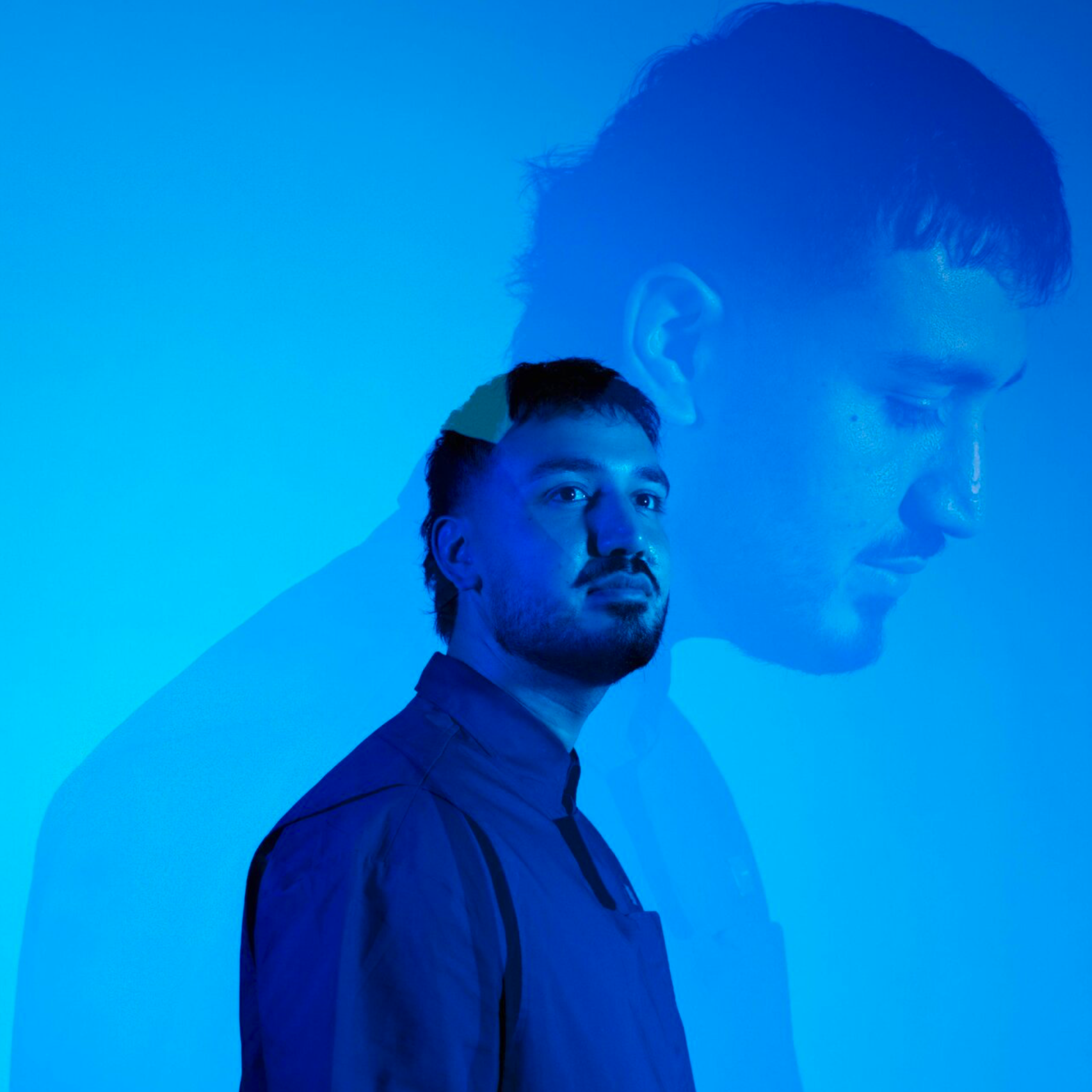 A man with dark hair and a beard standing in front of a wall with a large blue projection of his profile. The projection overlaps his face, creating a layered visual effect. Zuhair