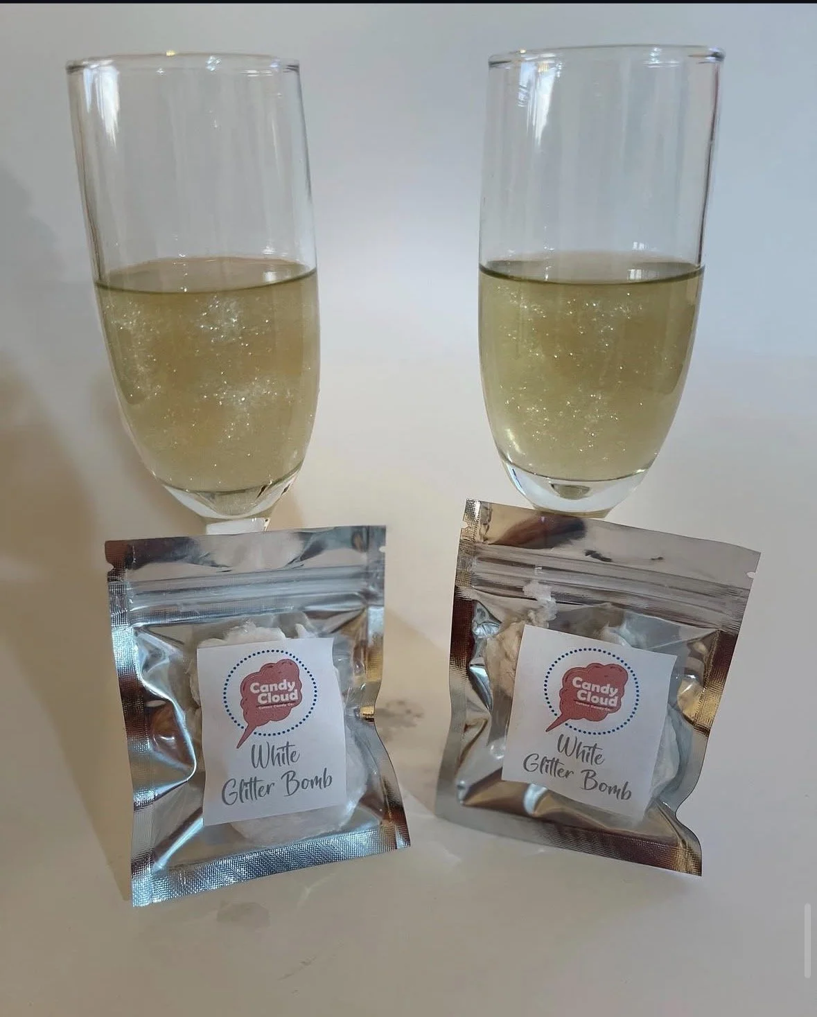Two champagne flutes filled with white sparkling beverage, each placed above a sealed bag labeled 'Candy Cloud White Glitter Bomb'.