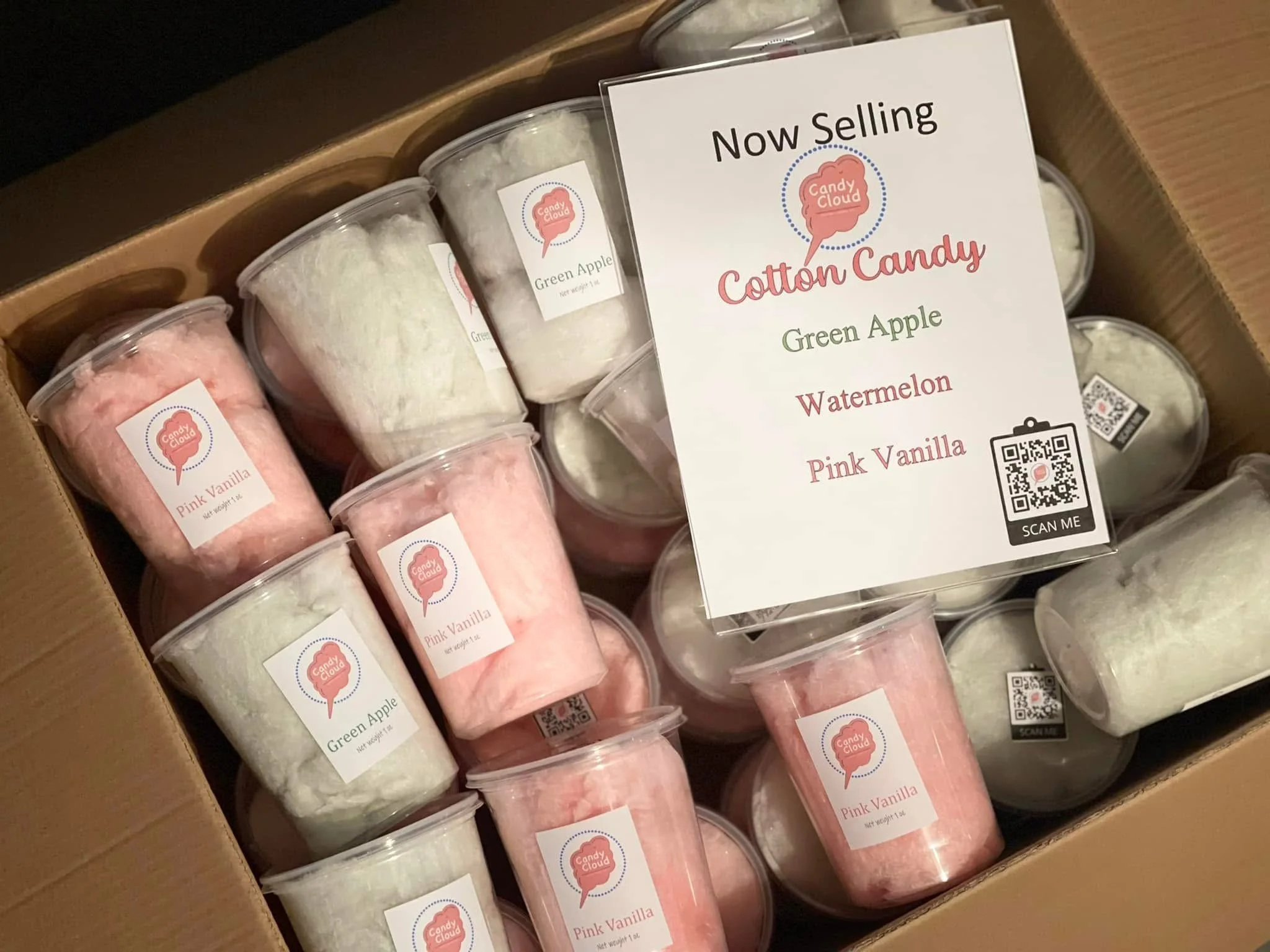 Box of cotton candy containers with pink vanilla, green apple, and white flavors, and a promotional sign listing flavors and a QR code.
