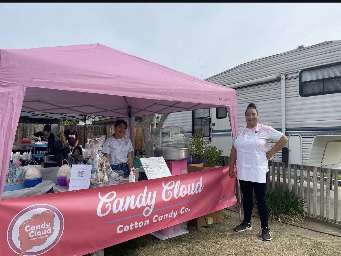 People manning a pink Candy Cloud Cotton Candy stand at an outdoor event with camper and RV in the background.