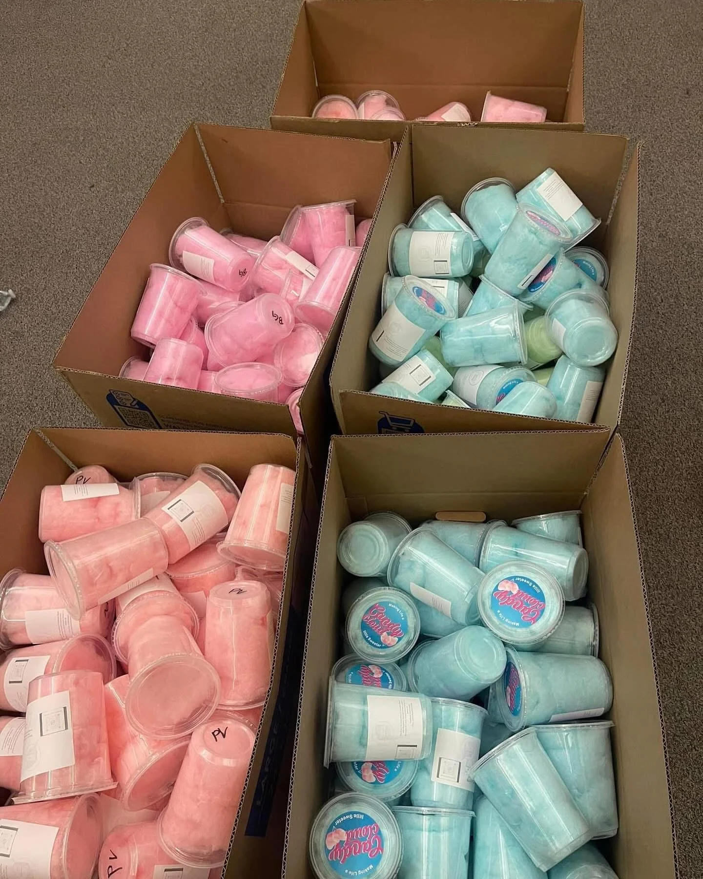 Four large cardboard boxes filled with pink and blue cotton candy containers on a brown carpeted floor.