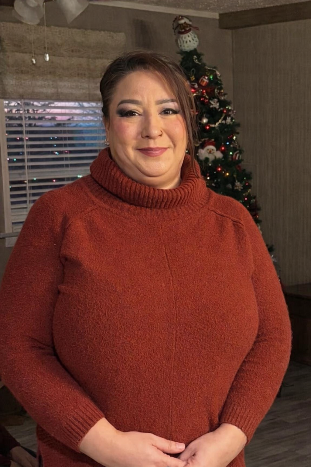A woman with short brown hair and makeup, wearing an orange turtleneck sweater, standing in a room decorated for Christmas with a Christmas tree in the background.