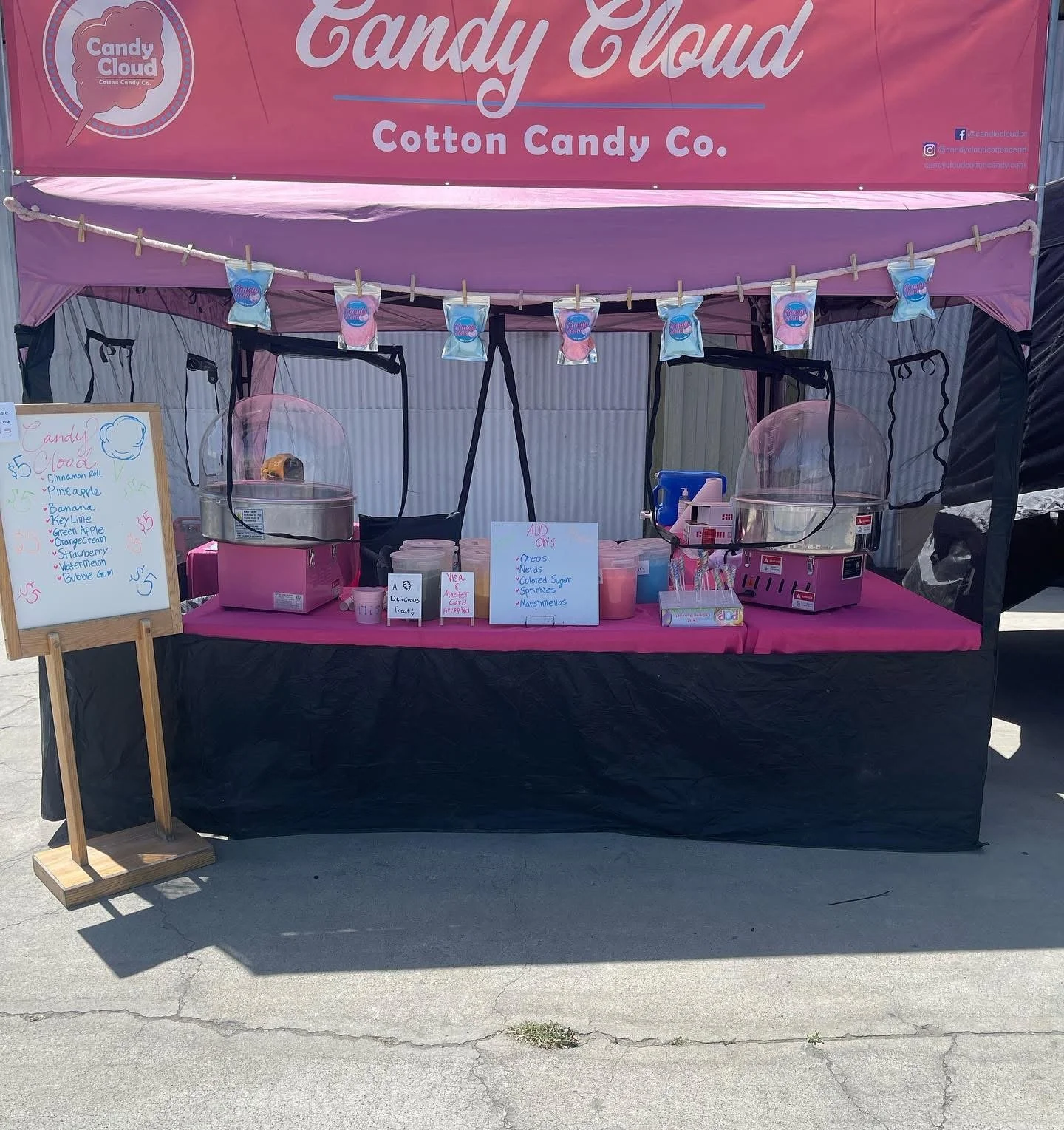 A pink and black outdoor cotton candy stand with a pink sign reading 'Candy Cloud' and 'Cotton Candy Co.' The stand has two cotton candy machines and a whiteboard listing different flavors and prices, with a row of small cotton candy bags hanging above.