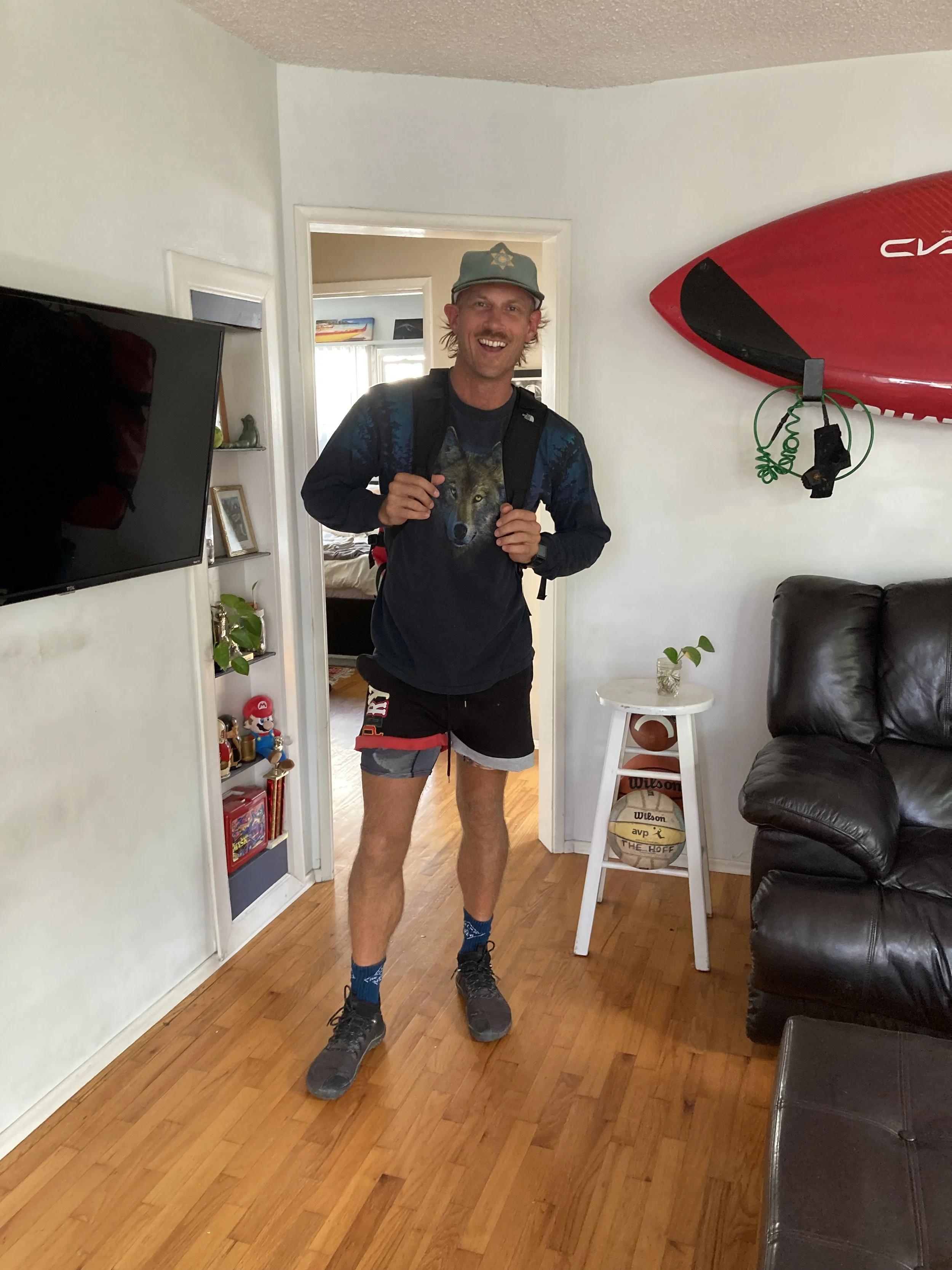 A man standing inside a living room, smiling, wearing a hat, a wolf-themed shirt, shorts, and hiking shoes, with a backpack.