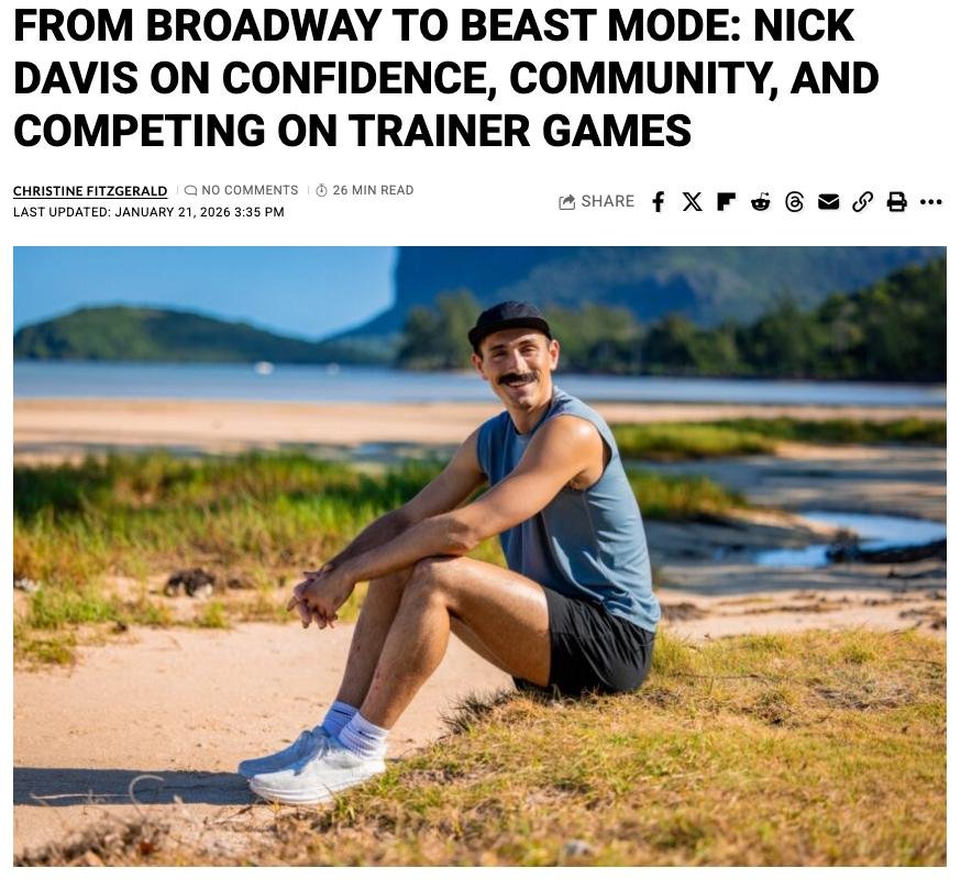 A man with a mustache, wearing a black cap, blue sleeveless shirt, black shorts, white socks, and sneakers, sitting on a sandy and grassy area near a body of water, with mountains and greenery in the background.