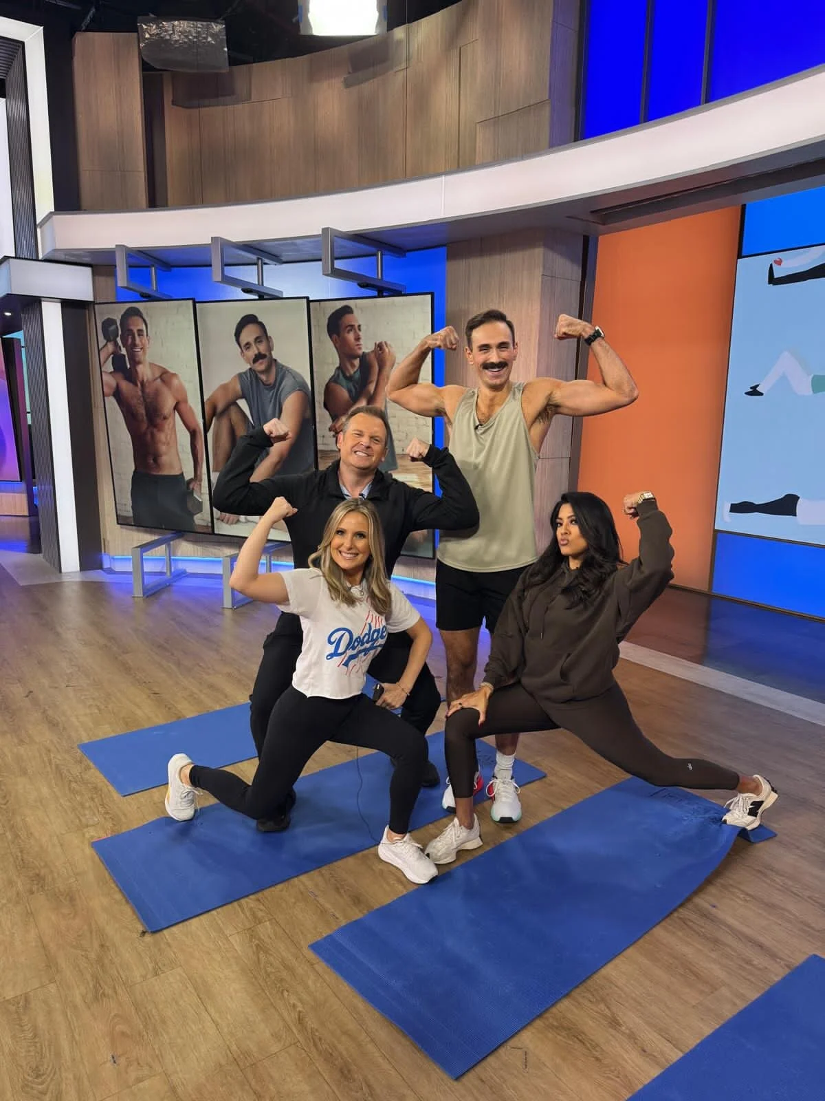 Four people in a gym studio posing with flexed muscles, with three posters of men in fitness poses hanging behind them. They are standing and kneeling on blue exercise mats, with large screens and monitors in the background.
