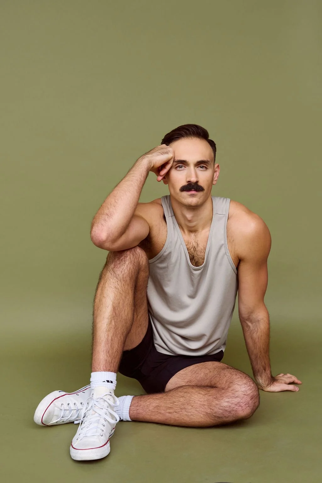 A man with a mustache, wearing a gray sleeveless shirt, black shorts, white sneakers, and white socks, sitting on the floor with his leg crossed, resting his elbow on his knee, against a plain green background.