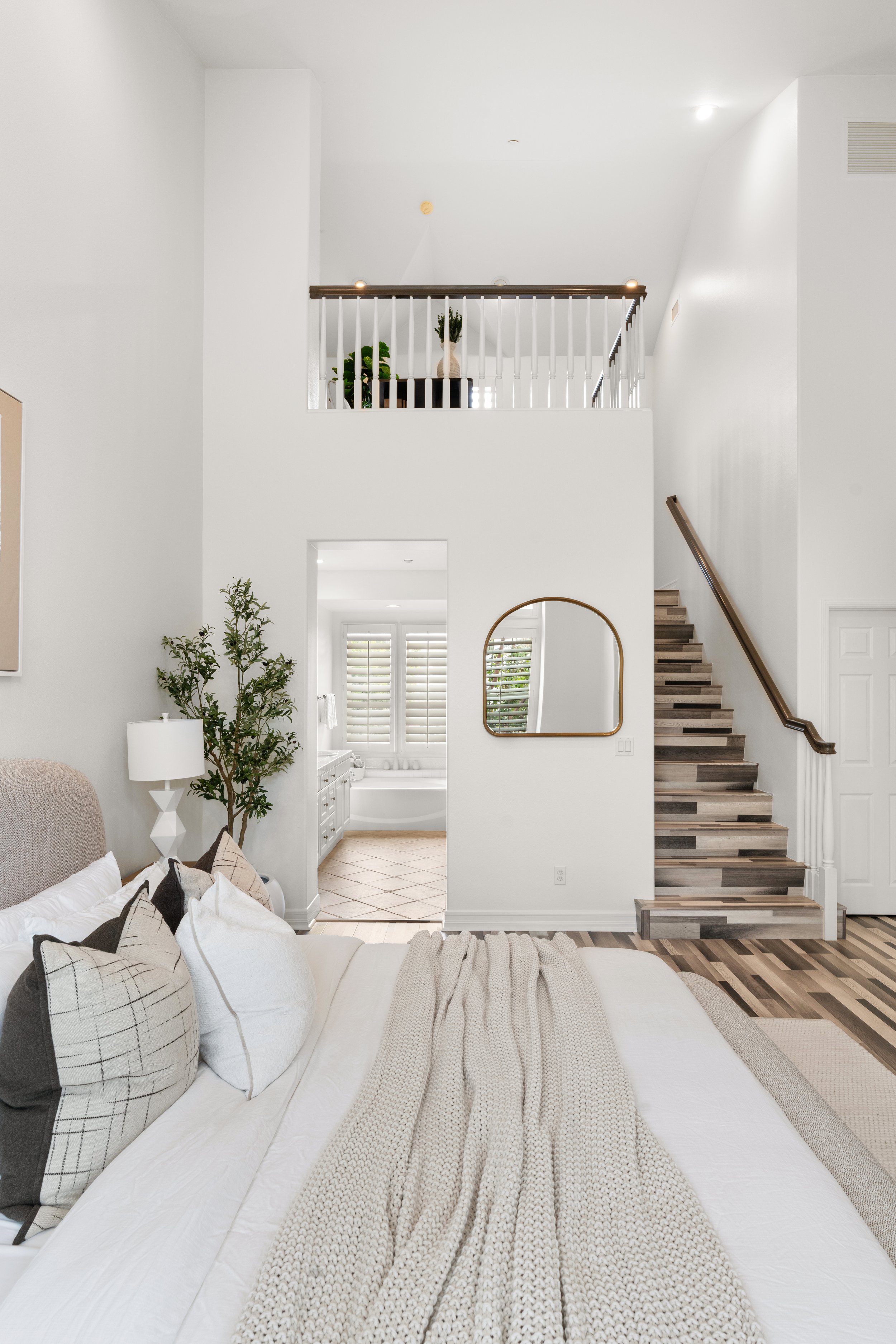 Interior view of a modern, minimalist bedroom with a staircase leading to an upper floor, a bedroom with a bathroom visible through an open doorway, and decorative elements including a mirror and plants.