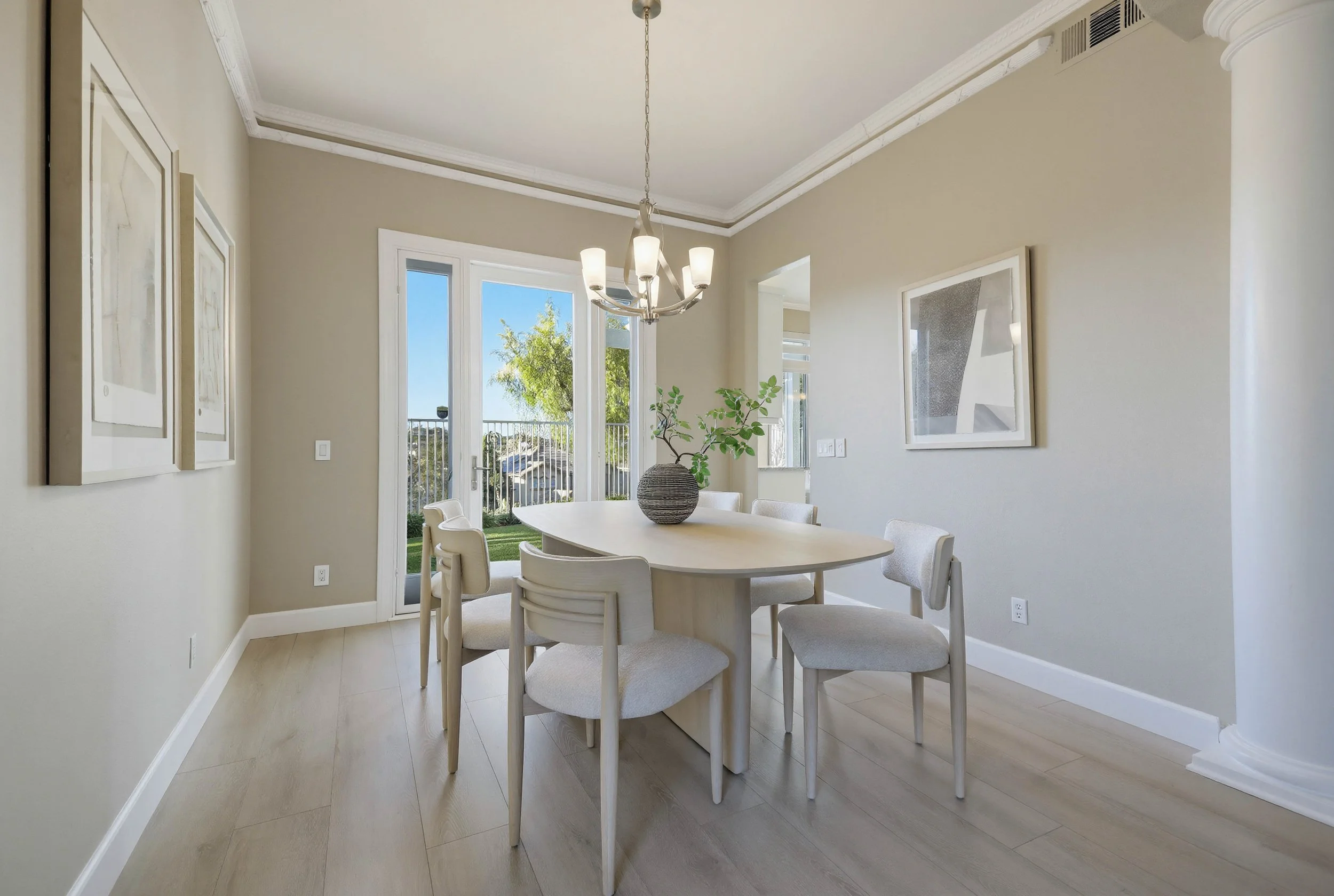 Dining room with a white oval table, six matching chairs, a vase with greenery, a chandelier, large glass door leading outside, light-colored walls, and framed artwork.