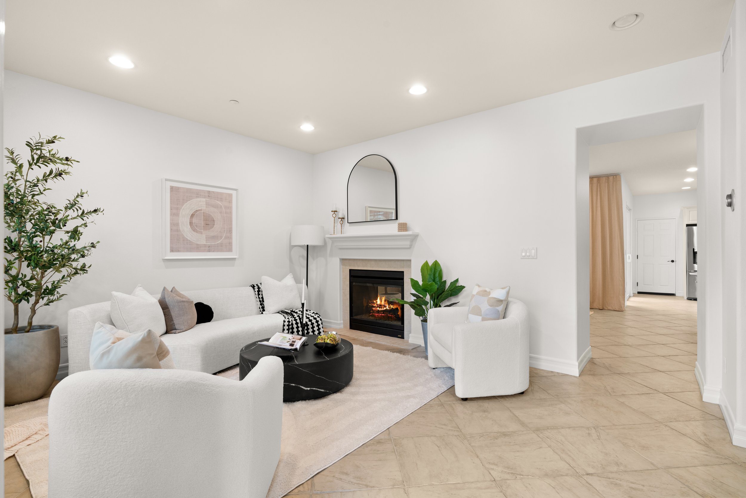 Living room with white sofa and armchair, fireplace with fire, coffee table, potted plant, wall art, and a hallway in the background.