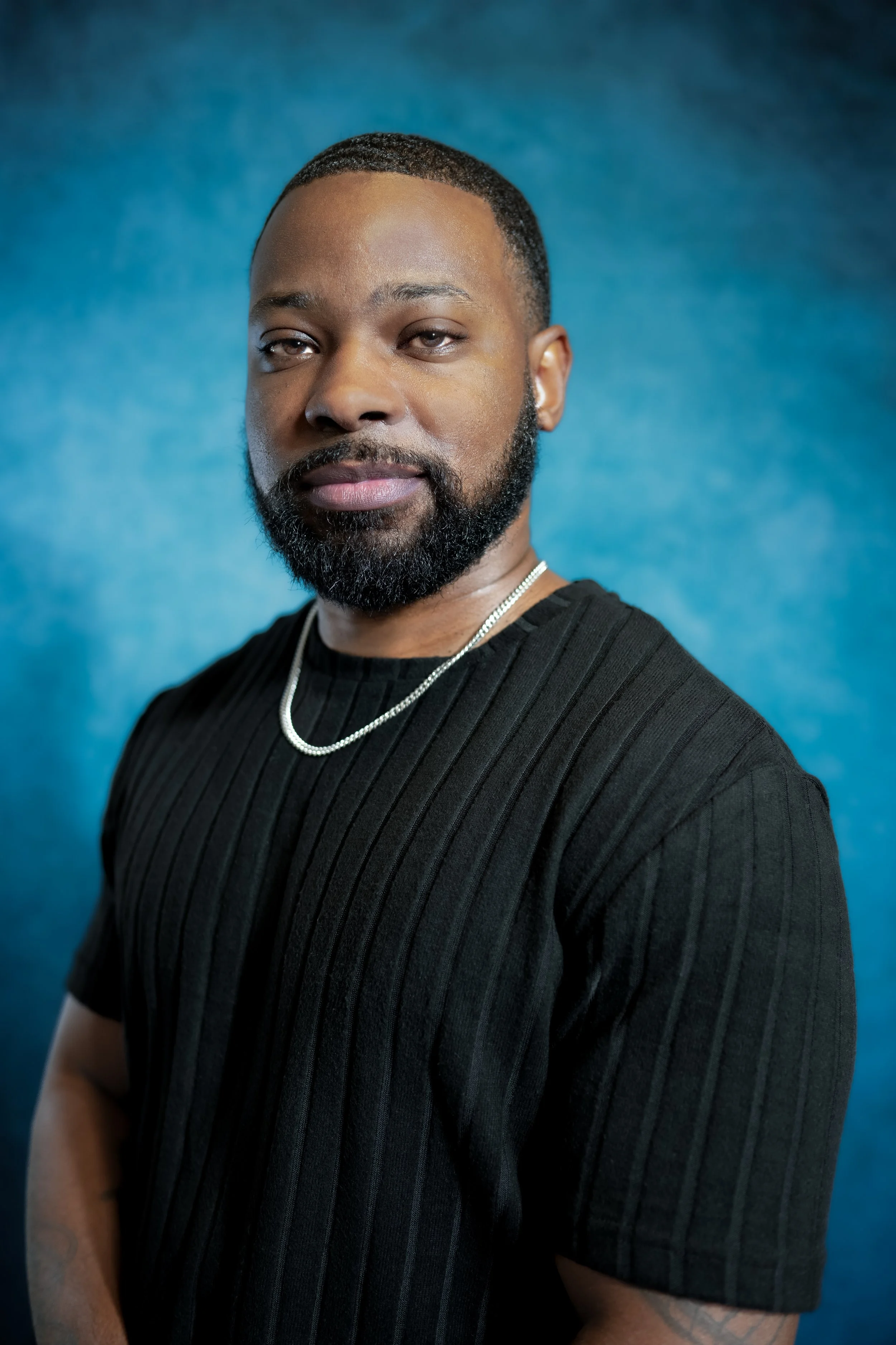 A man with a beard and short hair wearing a black ribbed shirt and a silver chain necklace stands against a blue background.