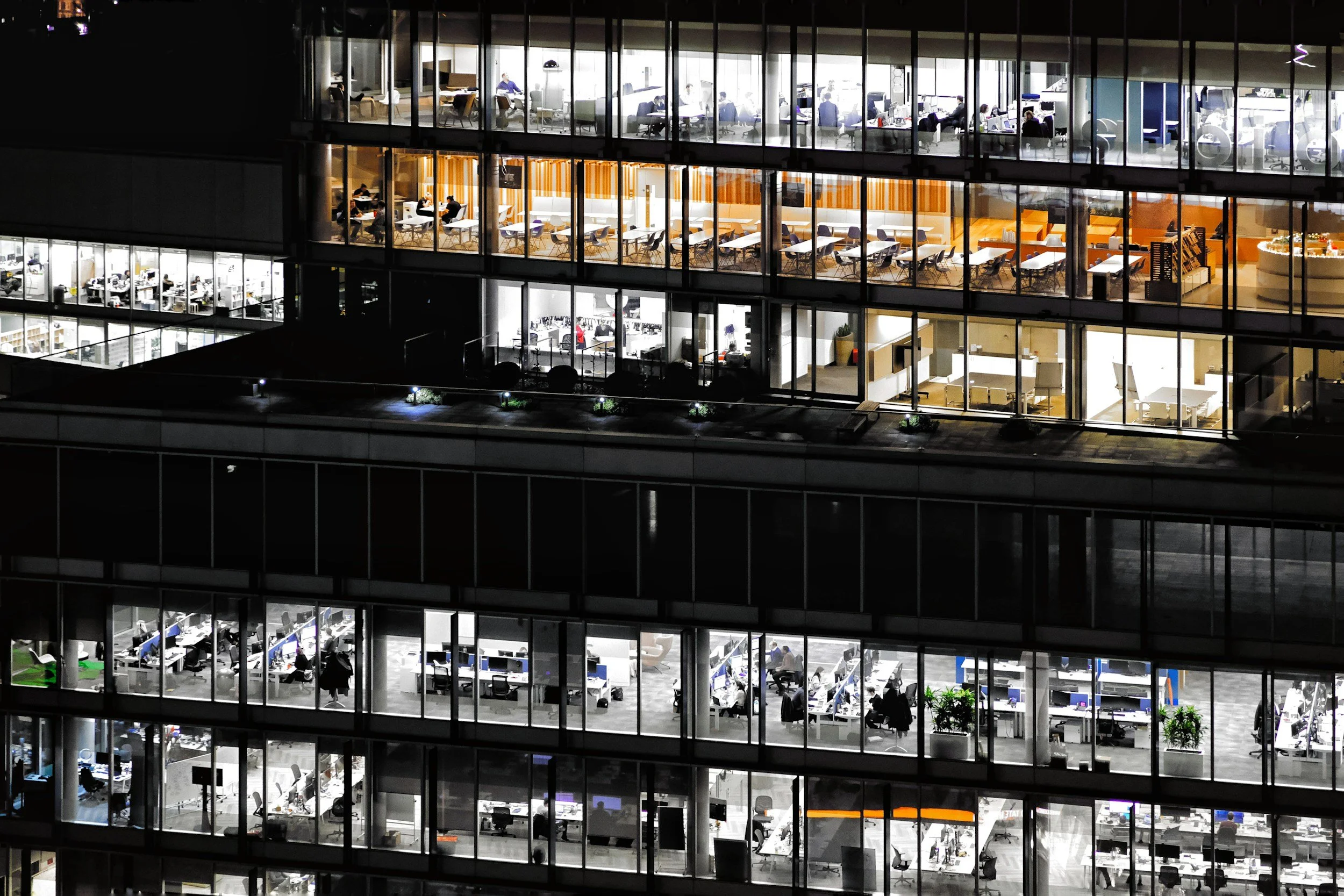 Nighttime view of a multi-story office building with illuminated floors. Each floor has different office setups with desks, chairs, and people working or sitting at tables, visible through large glass windows.