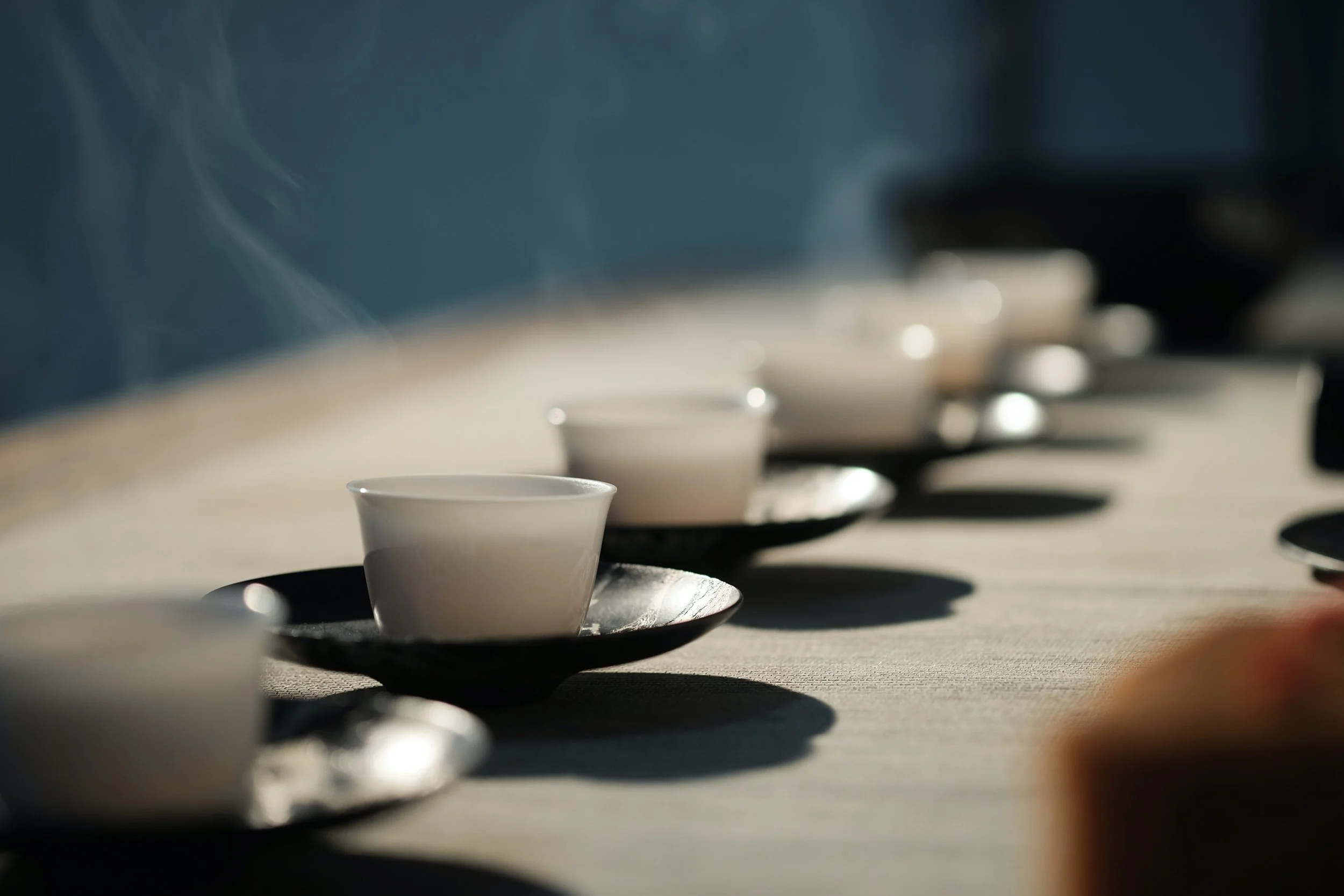 Several white cups on black saucers arranged in a line on a table with sunlight casting shadows. Tea meditation brings are deep connection to self, calmness and relaxation, and is a powerful form of meditation training.