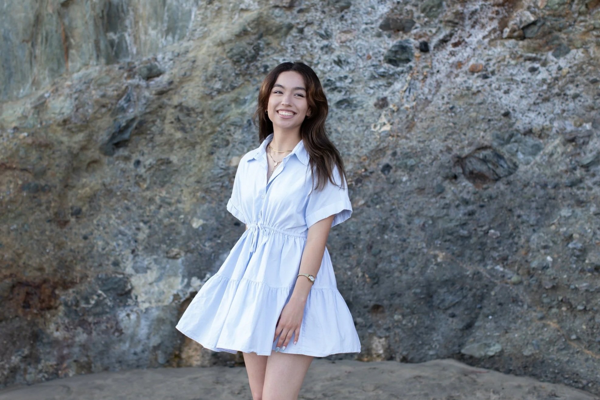A young woman with brown hair wearing a white dress and a watch, smiling at the camera against a rocky background.