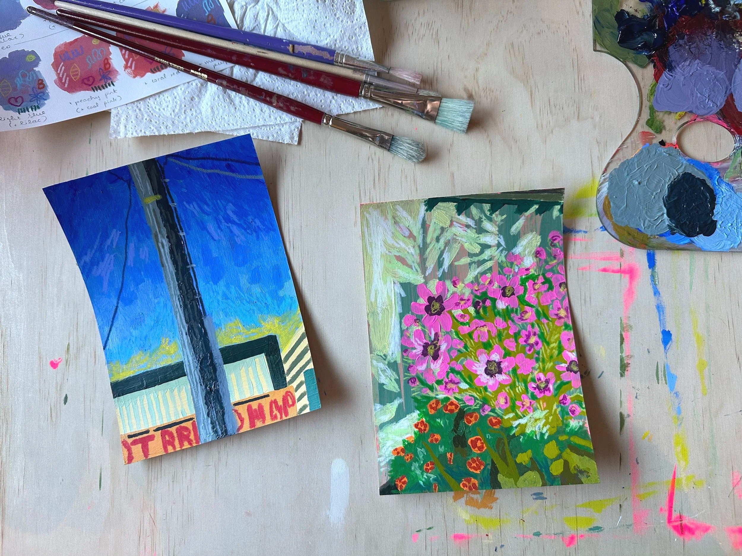 Photo of two painted artworks, one depicting 'Hot Bread Shop' and sky, and the other showing pink flowes in a garden. The artworks are placed on a wood surface, near paintbrushes and a paint palette.