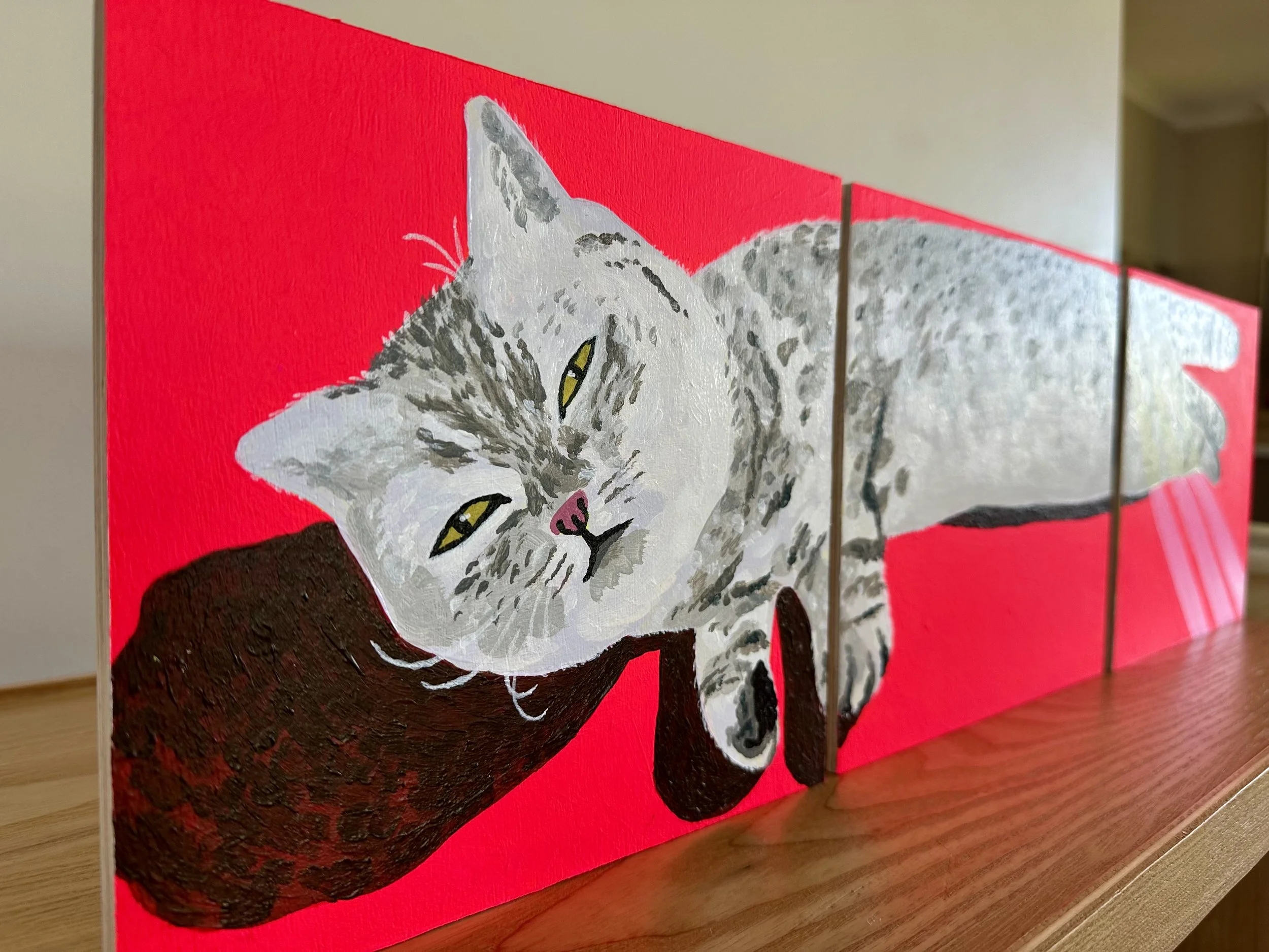 Photo of a painted tabby cat on a bright reddy pink background. The painting is in three parts, and sits atop a wood surface.