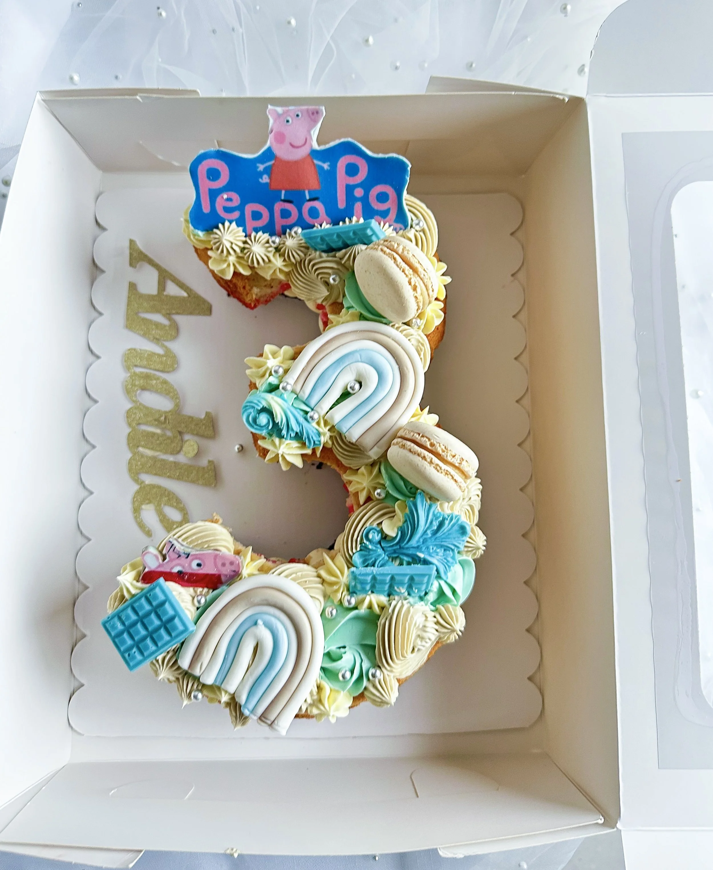 Number three birthday cake decorated with Rainbows, macarons, toys, and Peppa Pig-themed toppers, inside a white box.