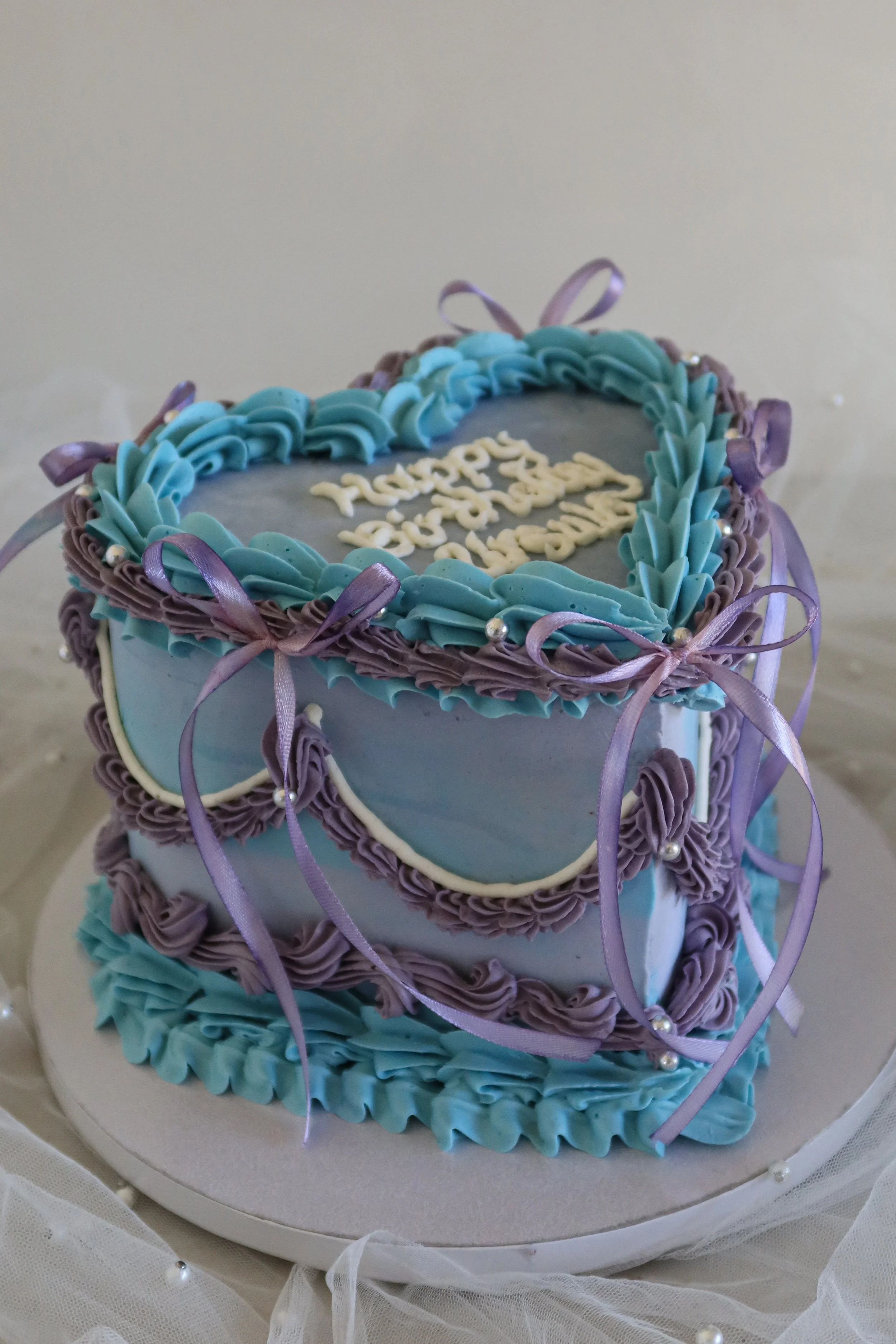 Decorative vintage heart birthday cake with blue and purple icing, ribbons, and pearl accents.