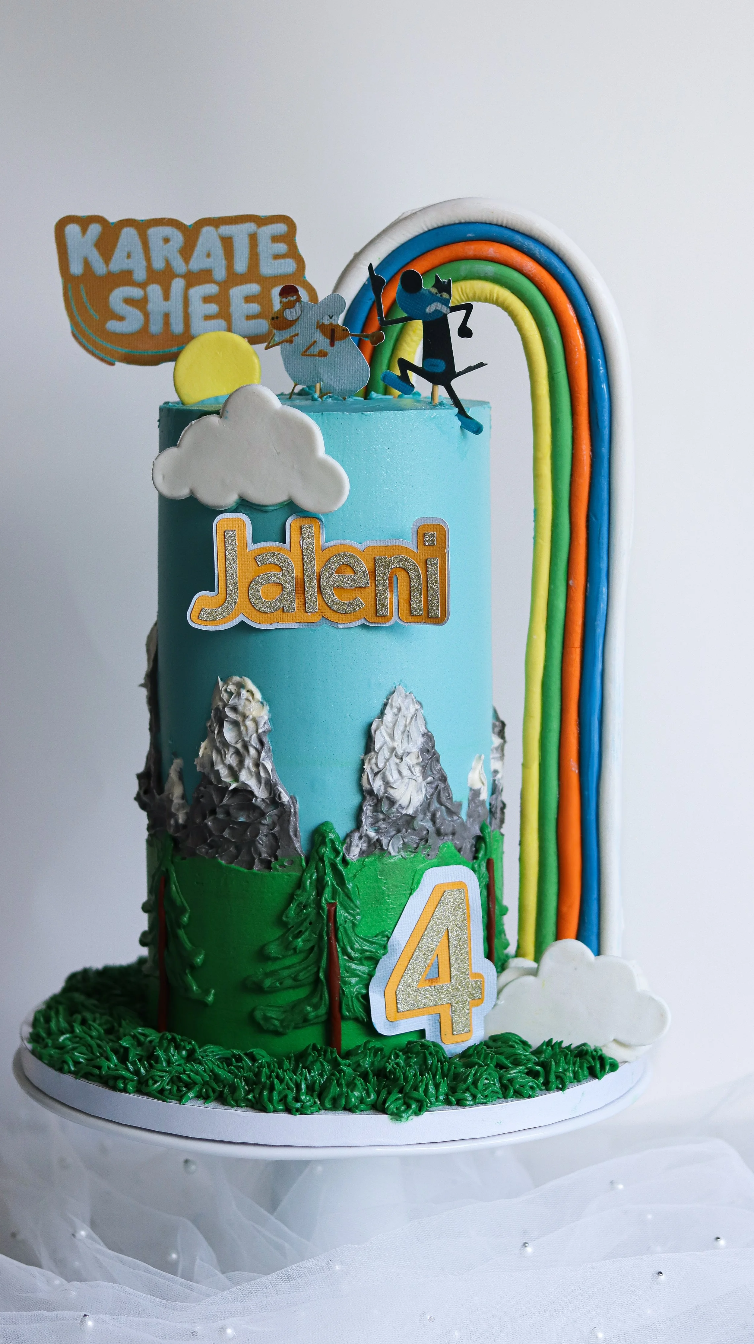 A colorful, multi-tiered birthday cake decorated with mountains, trees, clouds, a rainbow, and figures from the cartoon 'Kung Fu Panda.' The cake has the name 'Jaleni' and the number '4' on it, indicating a 4th birthday celebration.