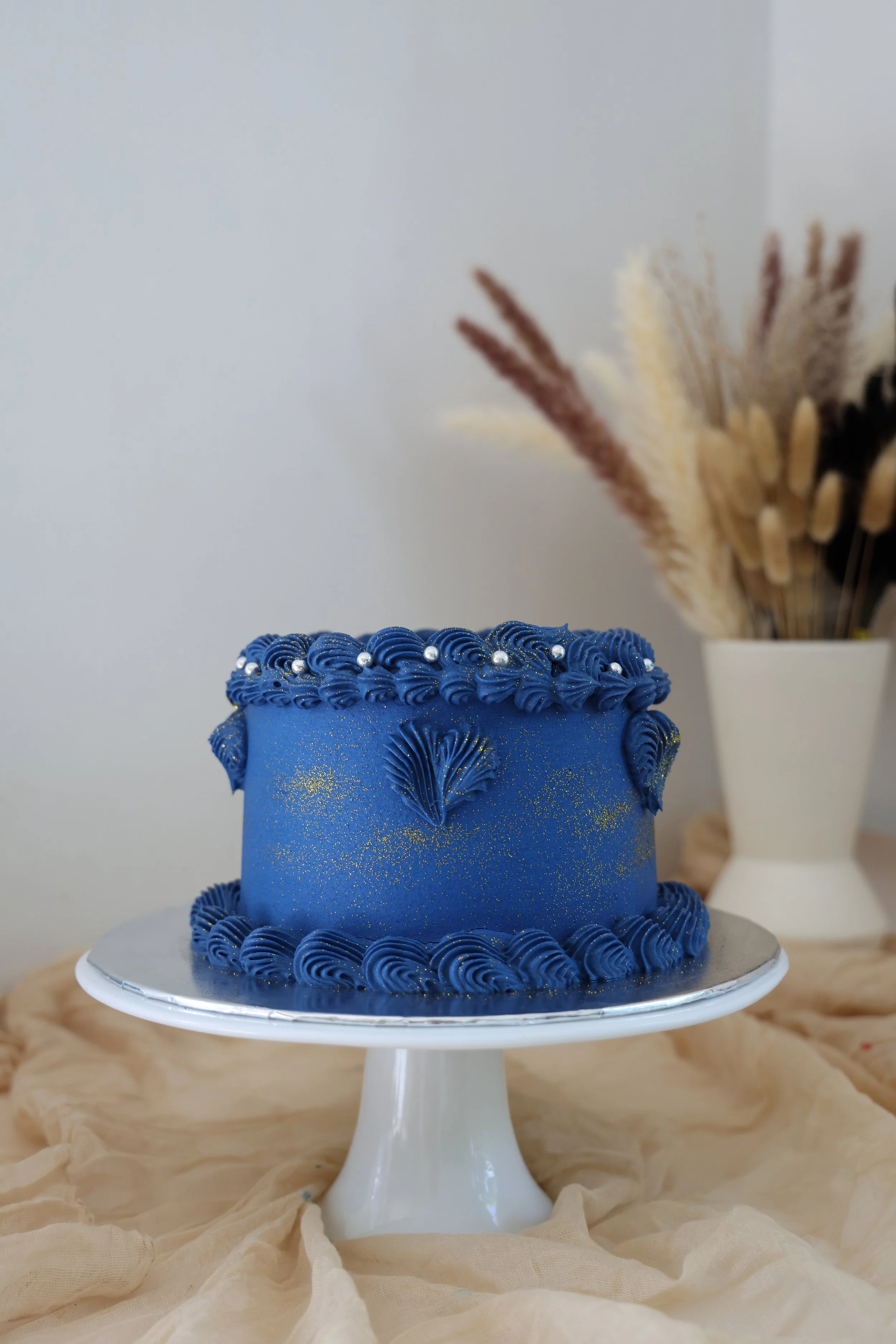 Blue decorated birthday cake with piped frosting and pearl accents on a white cake stand, with a vase of dried flowers in the background.