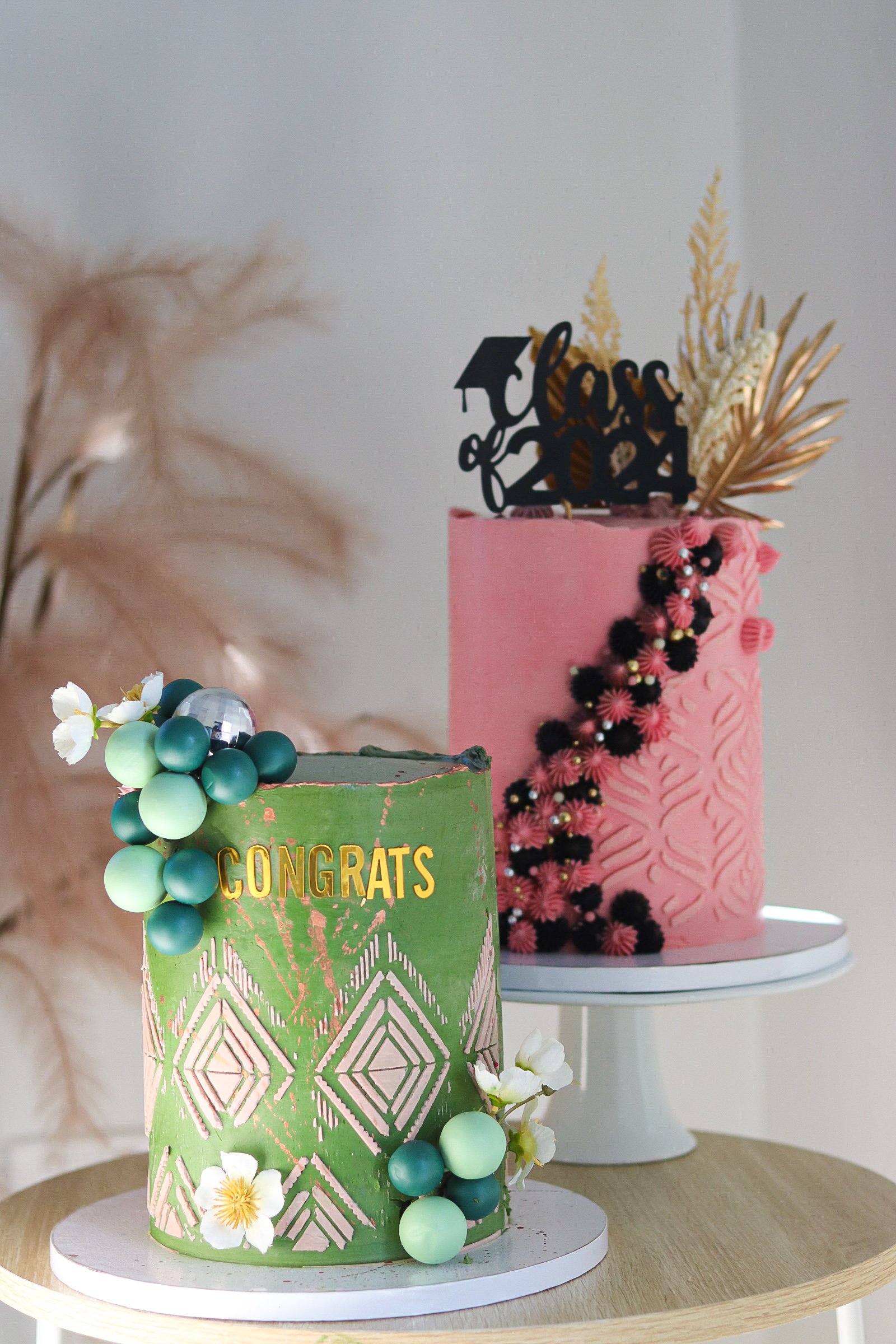 Two decorated celebration cakes, one green with the word "CONGRATS" and the other pink with "Class of 2024" topper, adorned with flowers, beads, and decorative leaves.