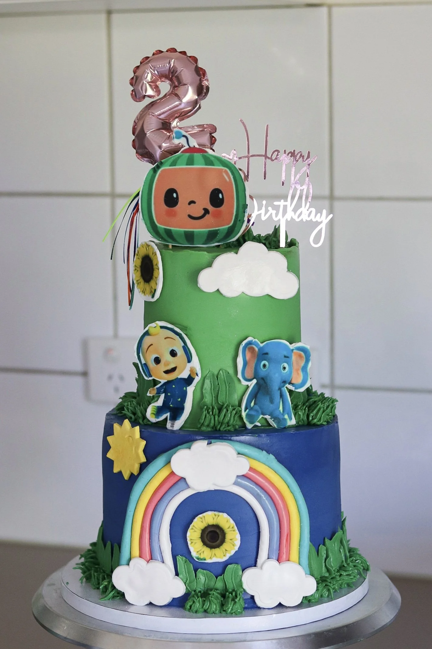 A three-tiered Cocomelon birthday cake decorated with characters from the show  Cocomelon characters. The bottom tier displays a rainbow with clouds and a sunflower. Green icing decorates the base of each tier. Cakes for kids.