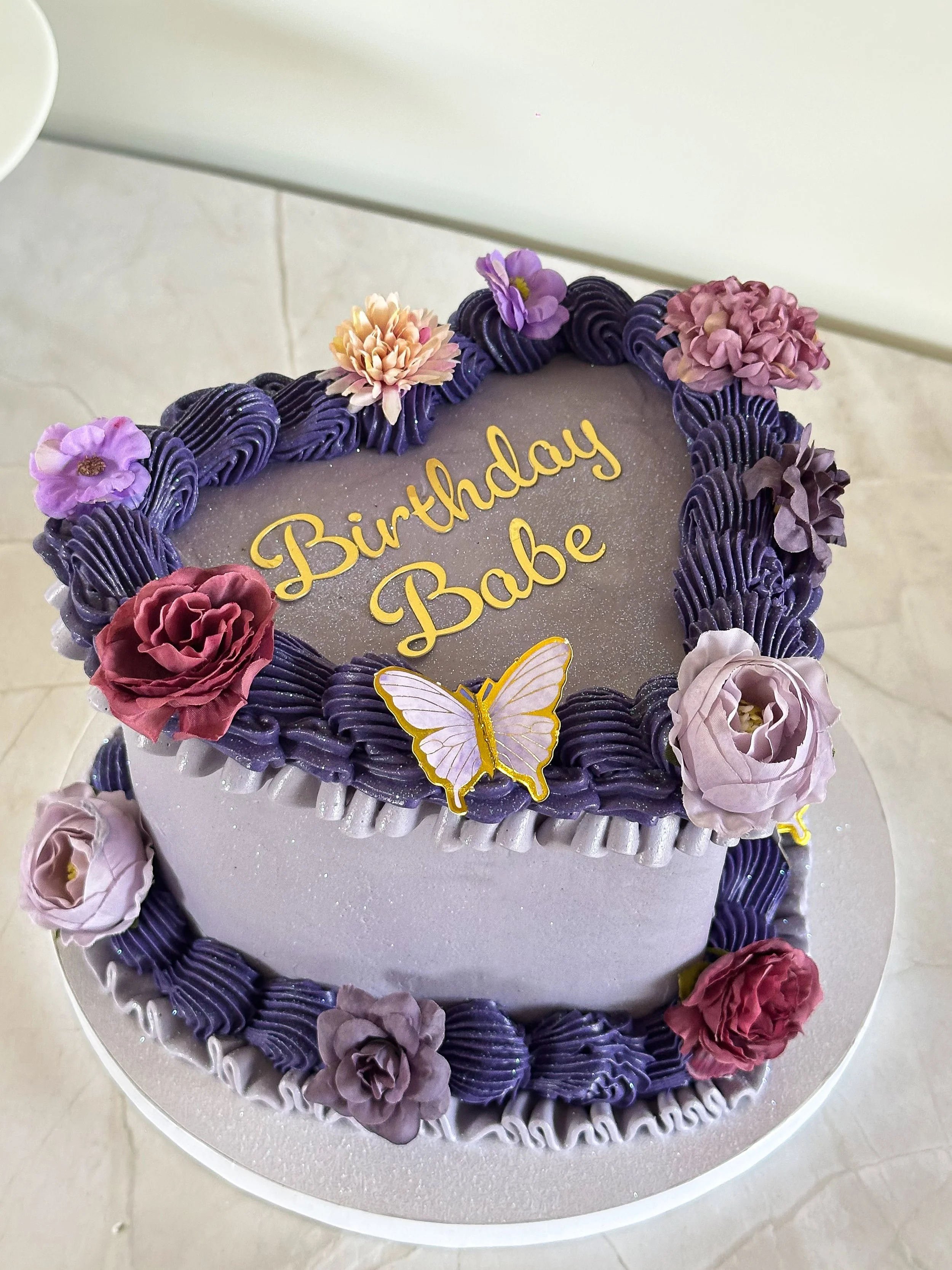 A heart-shaped birthday cake decorated with purple and pink flowers, purple icing borders, and a 'Birthday Babe' message with a butterfly decoration.