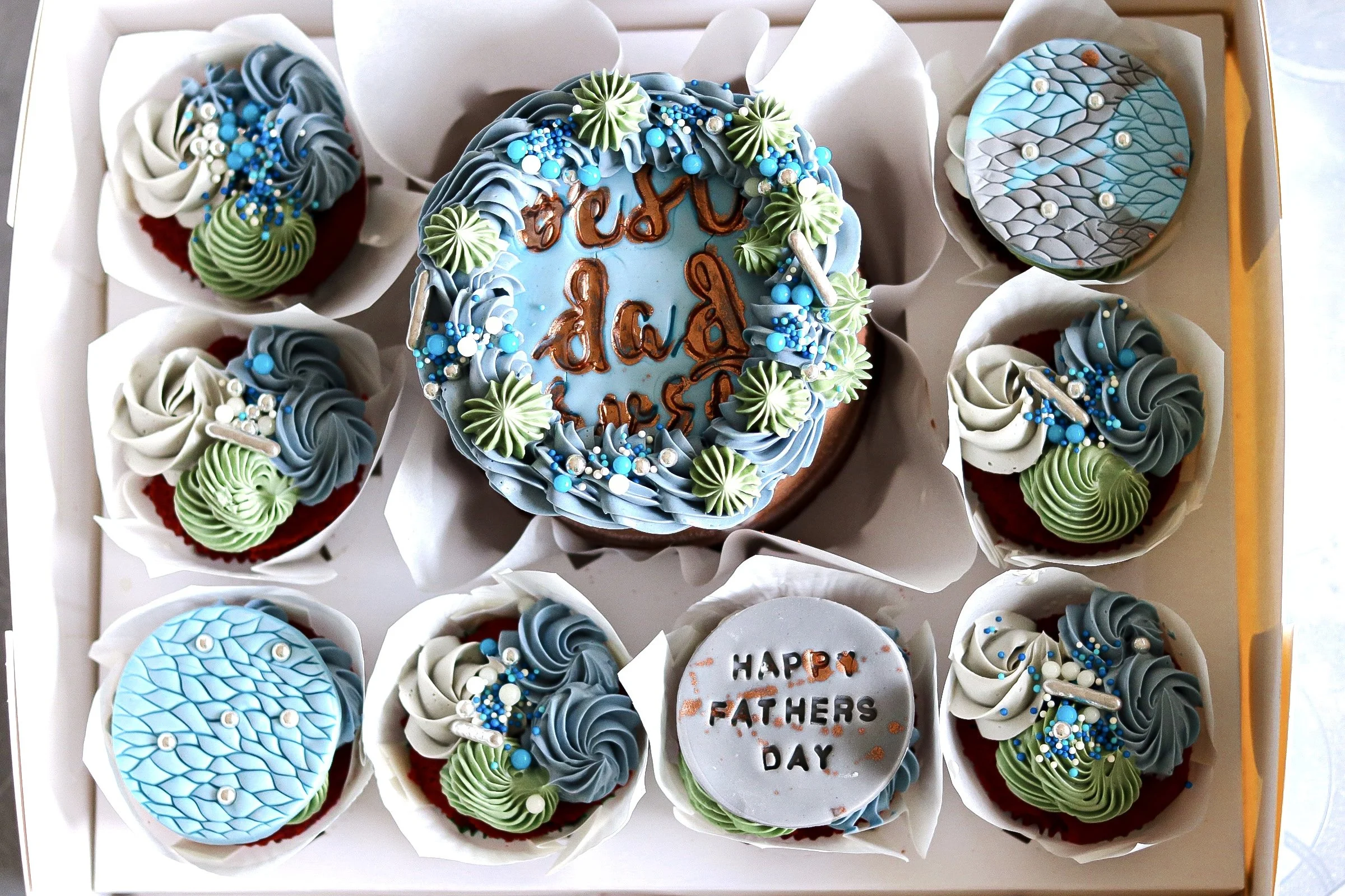 A Father's Day bento cake and cupcakes  with blue icing and decorative green, white, and blue fondant toppers surrounded by cupcakes with similar decorations and message cookies.