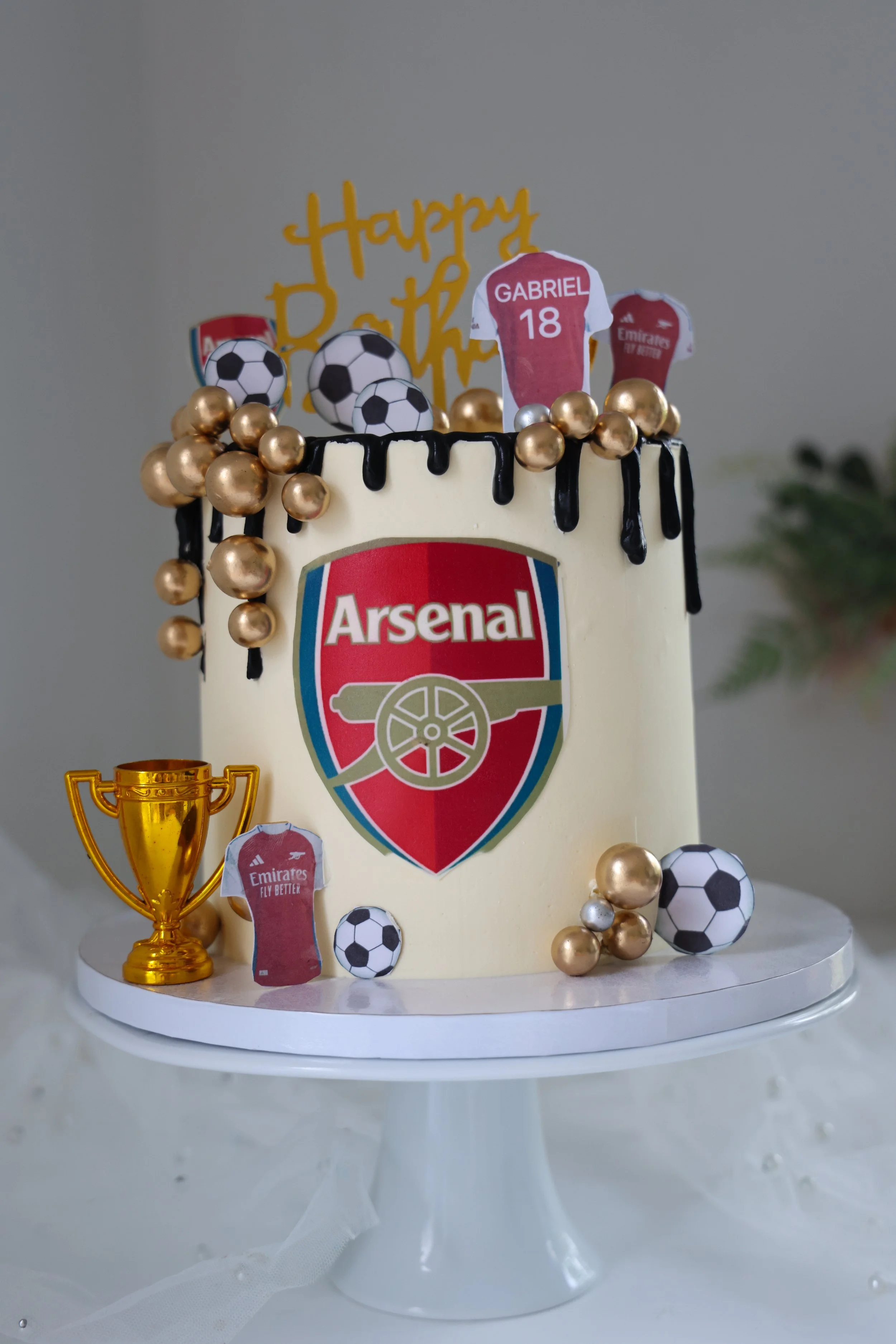 Football-themed decorated cake with Arsenal logo, soccer ball decorations, a gold trophy, and birthday elements on a white cake stand. Soccer themed cake.