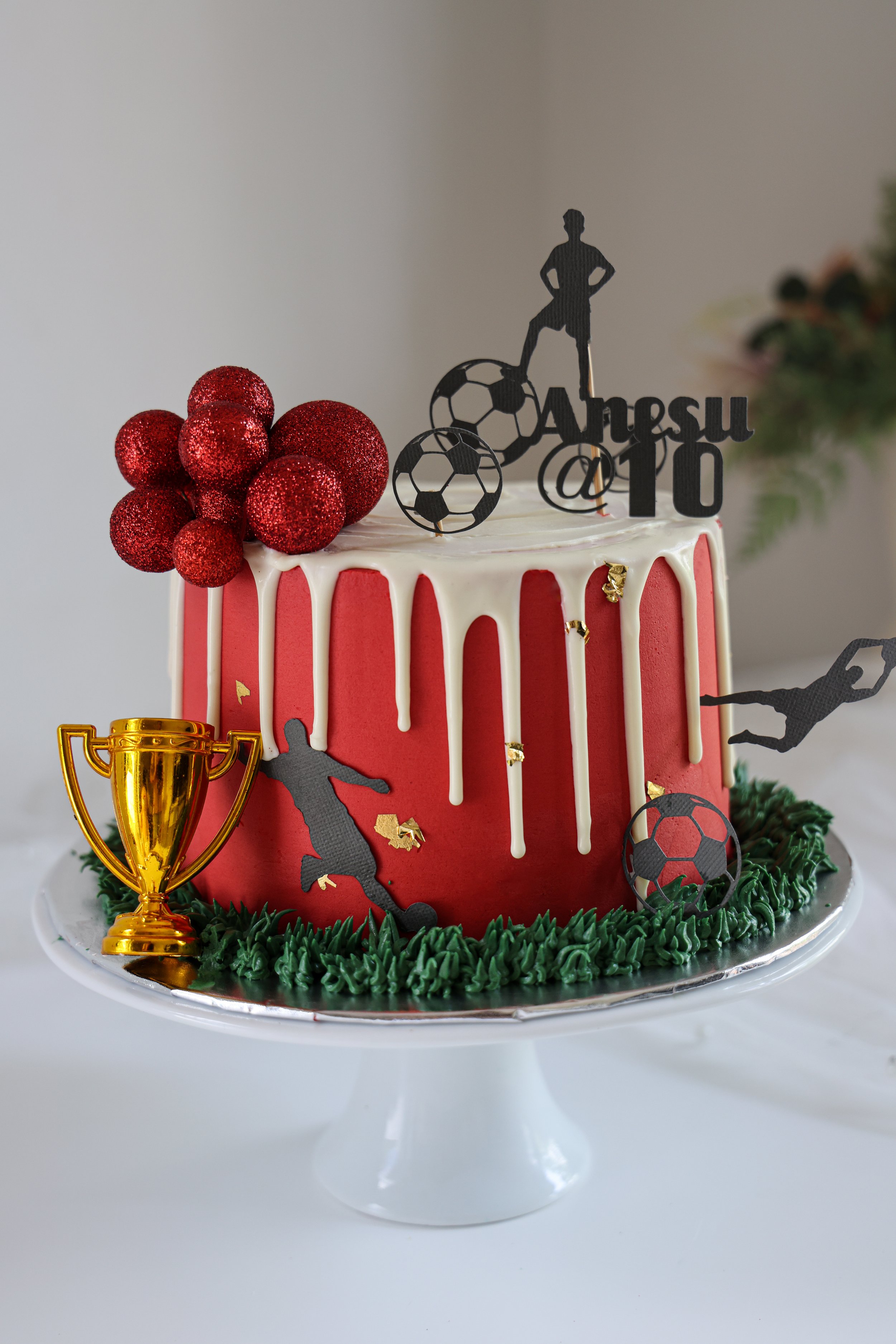 A red and white soccer-themed birthday cake decorated with figurines, a trophy, red glittery berries, and gold accents on a white cake stand.