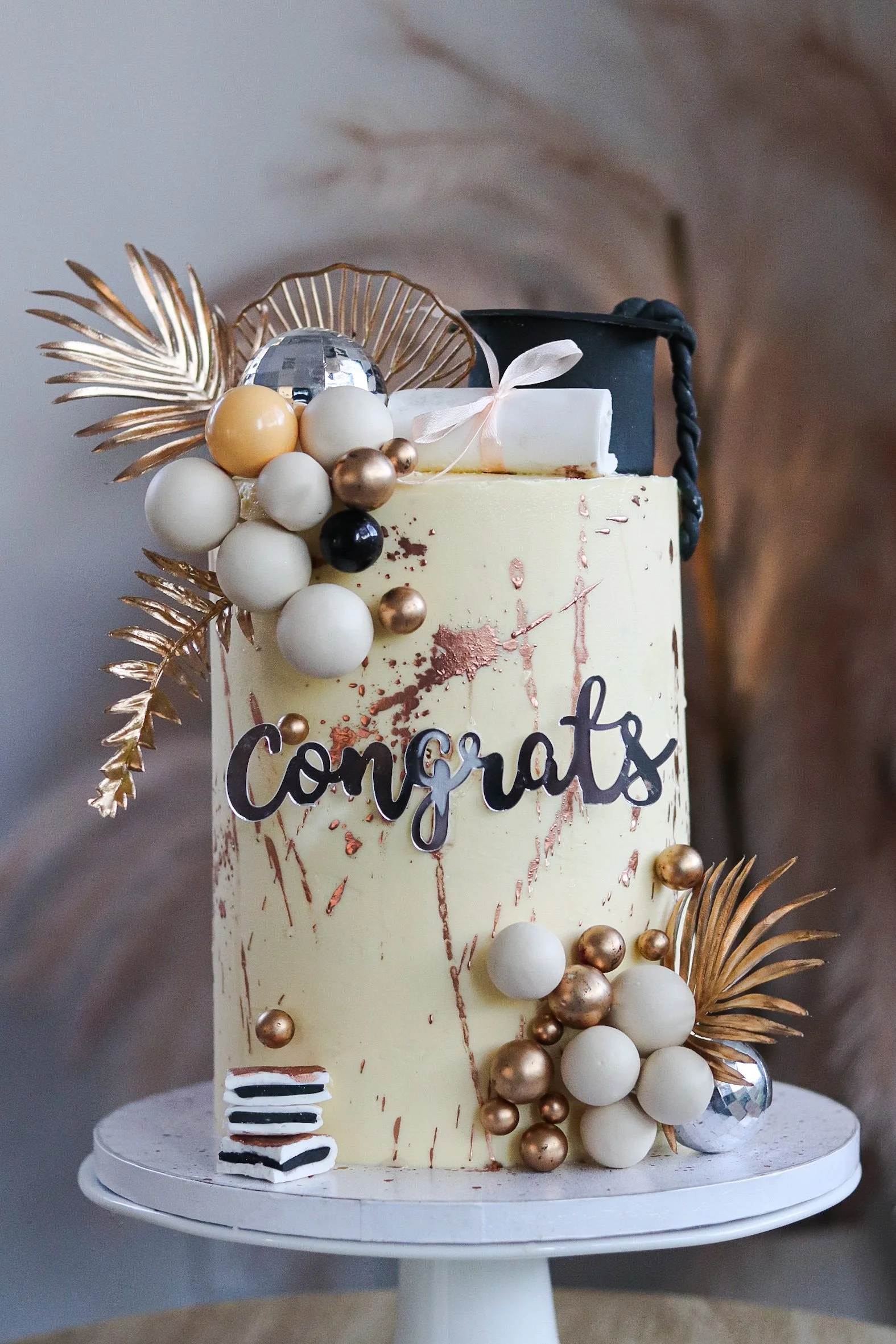 A decorated birthday cake with a "Congrats" sign, gold and white ornaments, gold leaves, a rolled document, and a small slice of cake on the base.