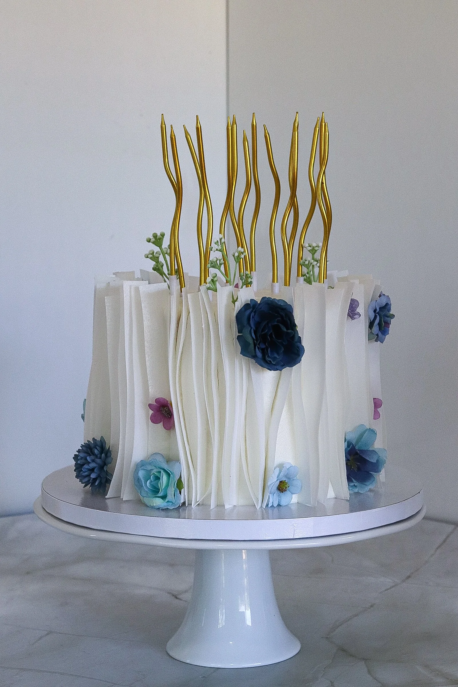 A multi-layered white elegant cake decorated with blue and purple flowers, topped with golden, twisted candle holders.