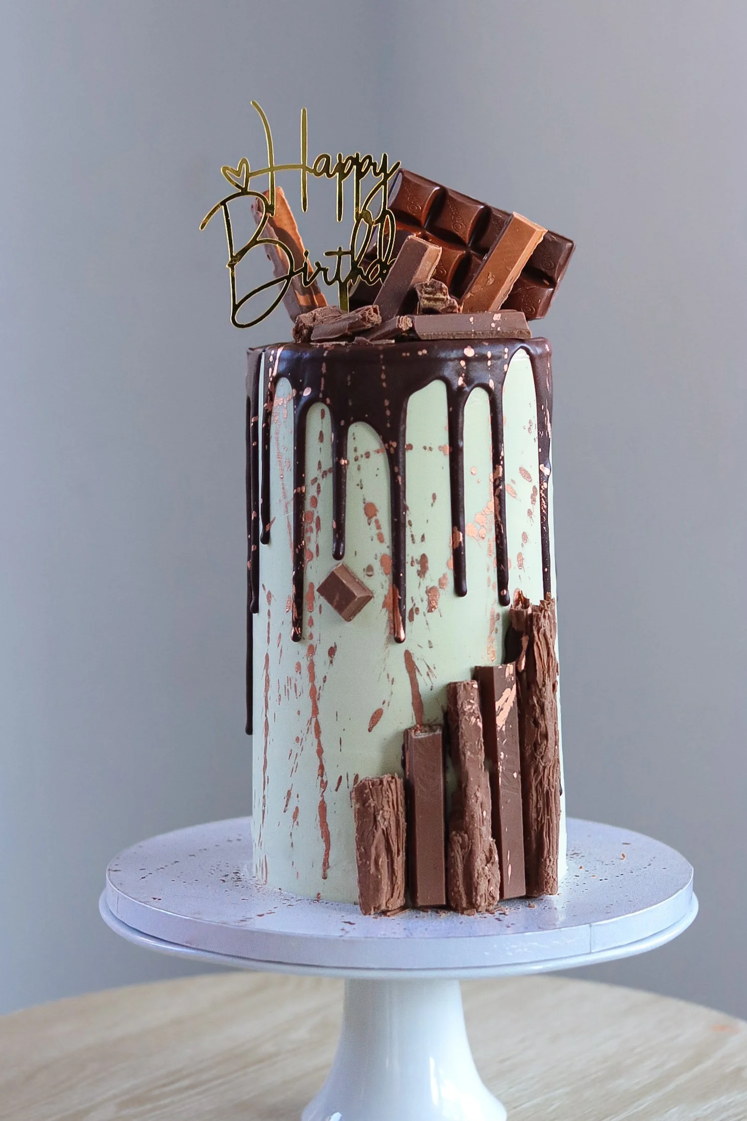 A tall birthday cake with white and green marbled icing, topped with a 'Happy Birthday' gold cake topper, chocolate drizzle, assorted chocolate bars, and chocolate pieces, displayed on a white cake stand.