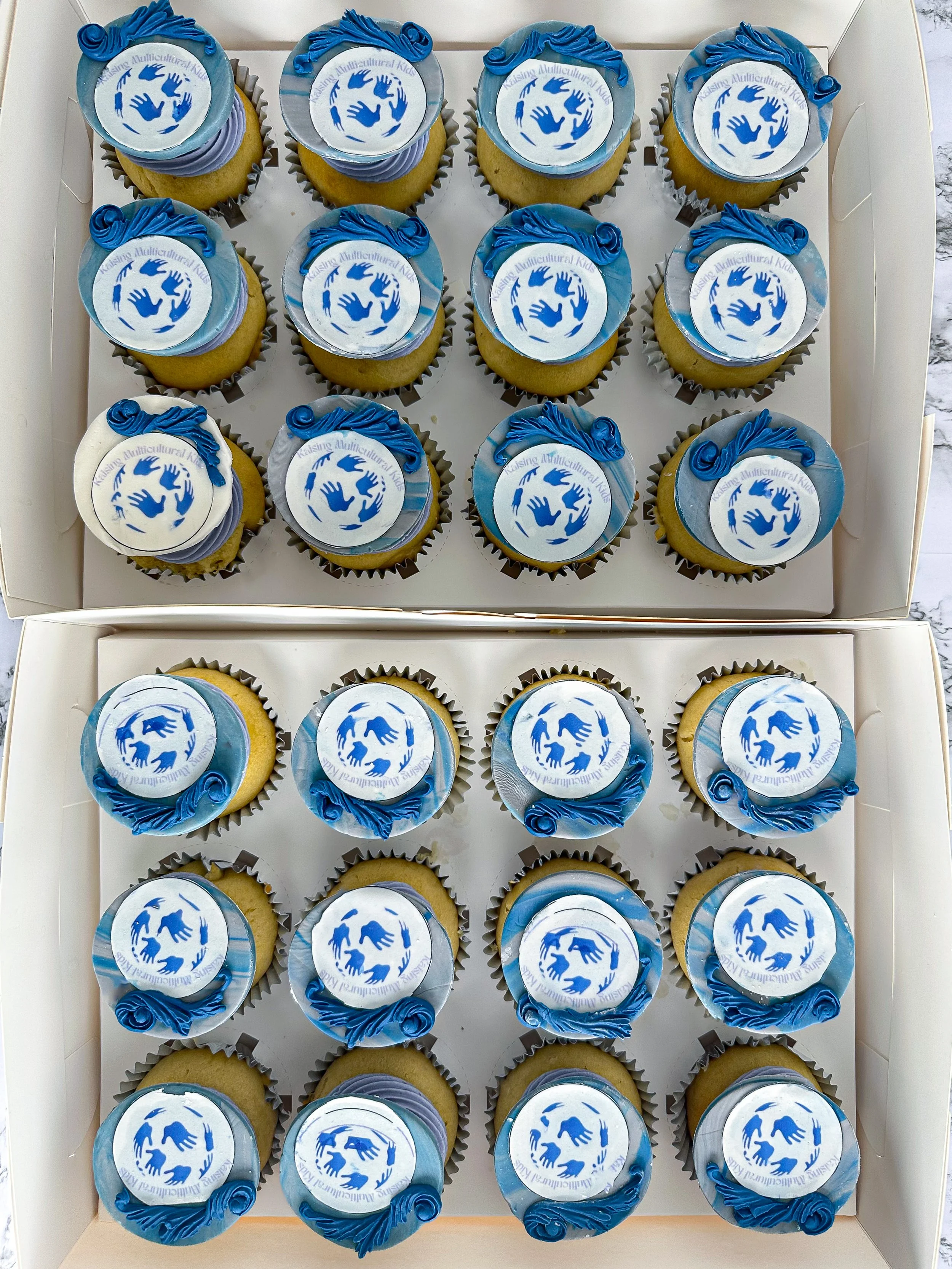 Two boxes filled with corporate cupcakes decorated with white and blue icing, featuring a circular logo with blue hands in a circle and text around it, and topped with blue icing decorations.