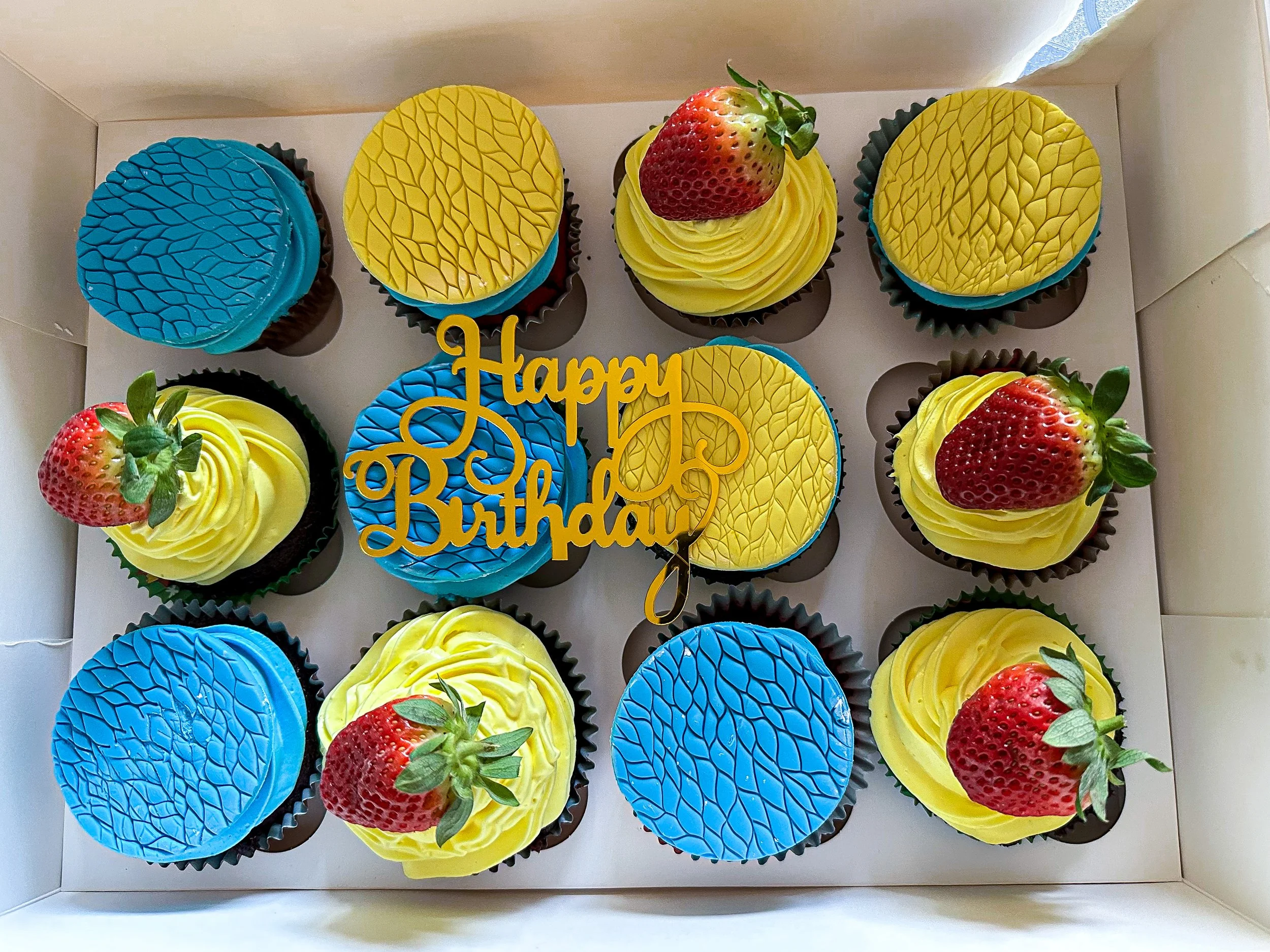 A box of birthday cupcakes decorated with blue and yellow icing, topped with fresh strawberries, and a yellow 'Happy Birthday' topper in the center.